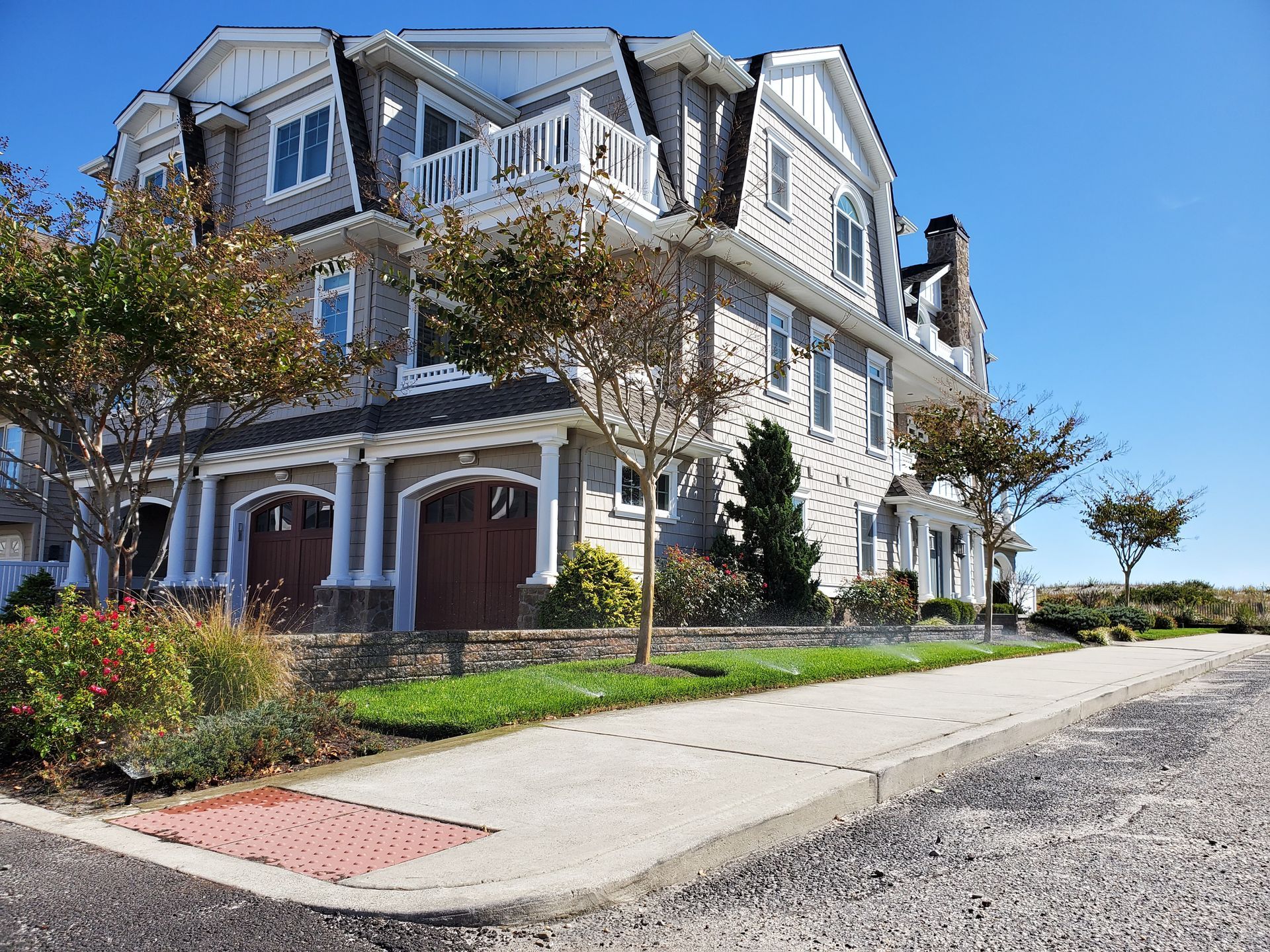 Three-story gray house with brown garage doors, trees, and a sidewalk on a sunny day.