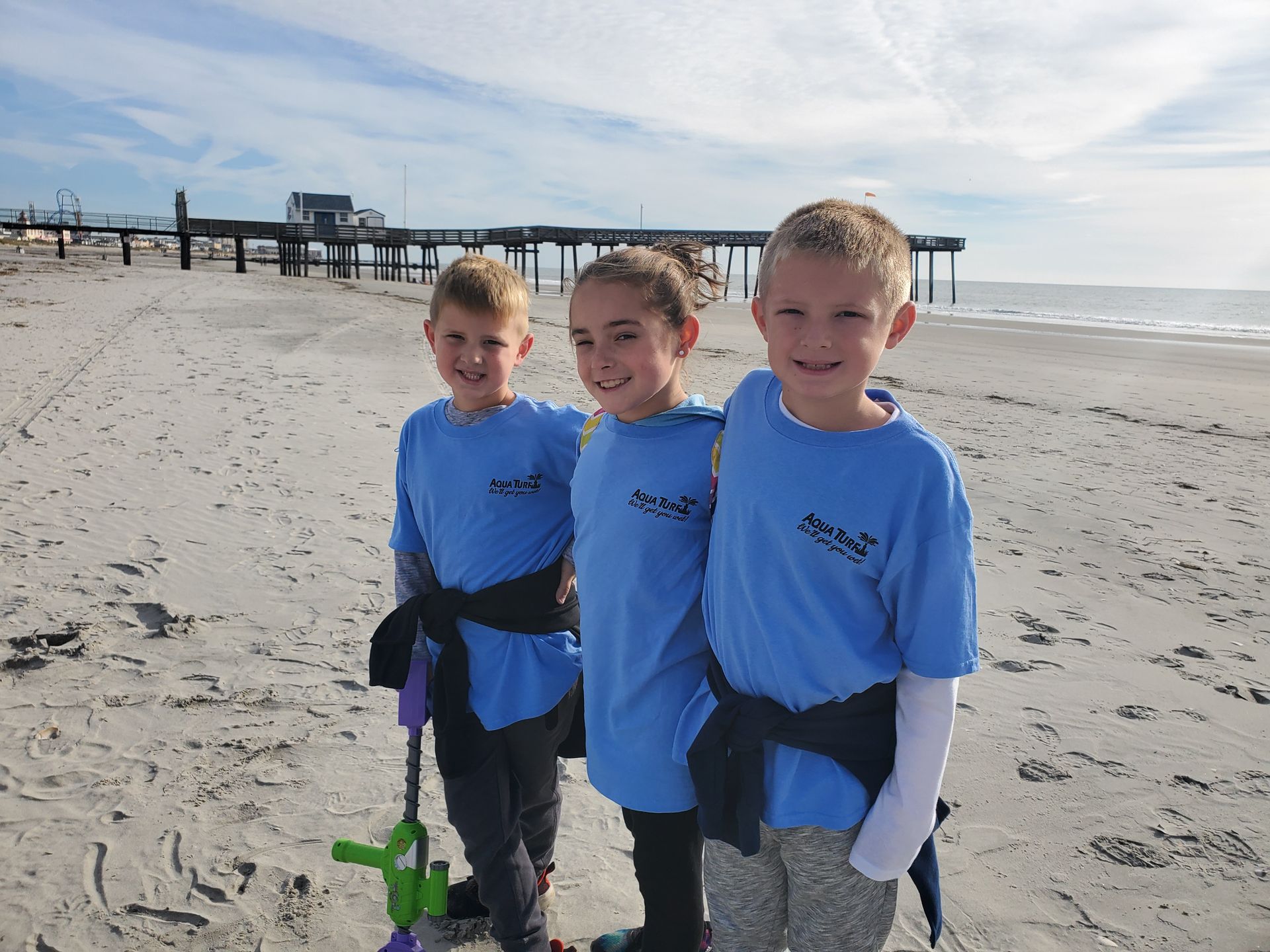 Three children in blue shirts and pants on a sandy beach, posing with an ocean pier in the background.