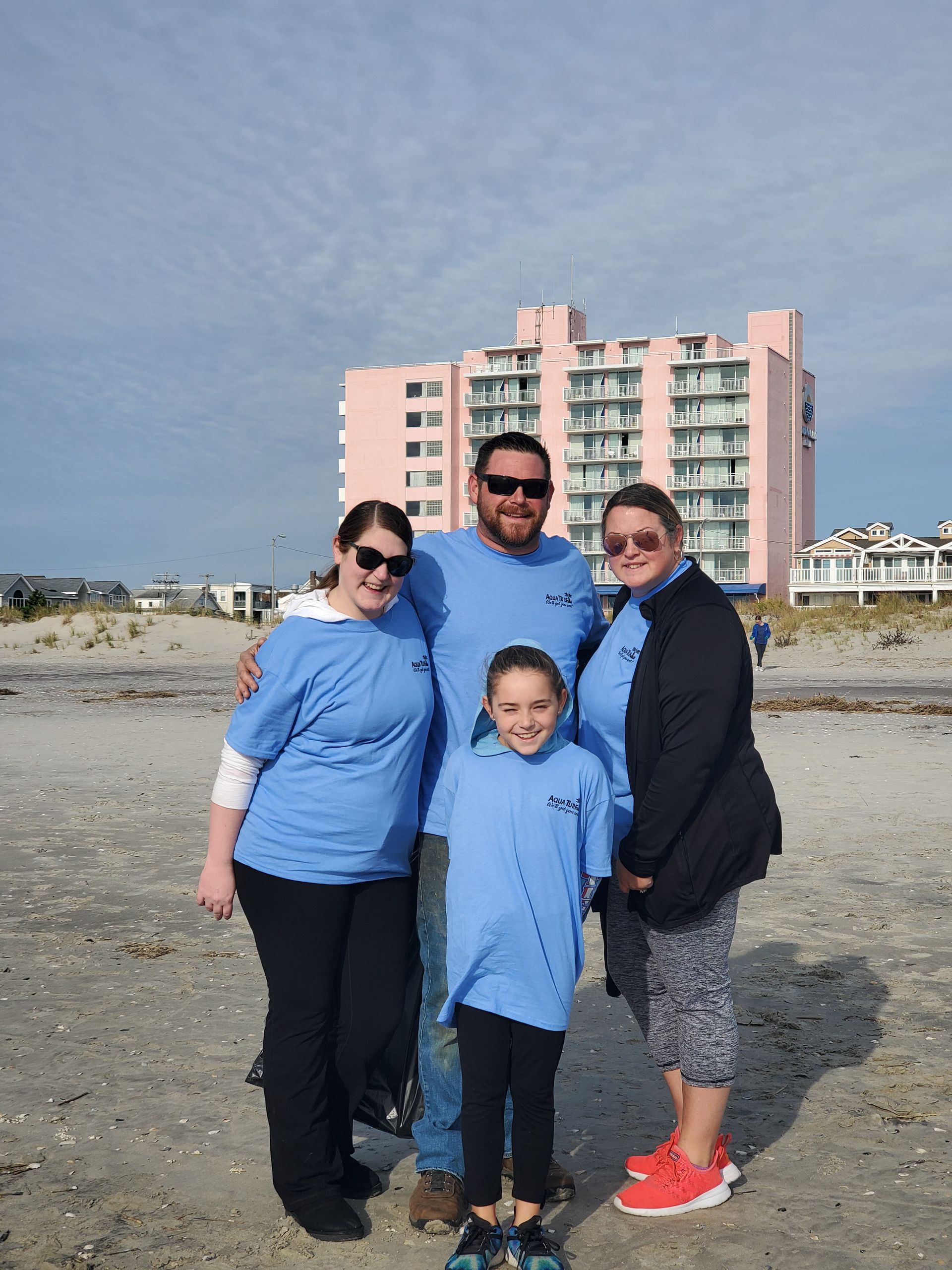 Family of four on a beach, wearing blue shirts, with a pink building in the background. Cloudy sky.