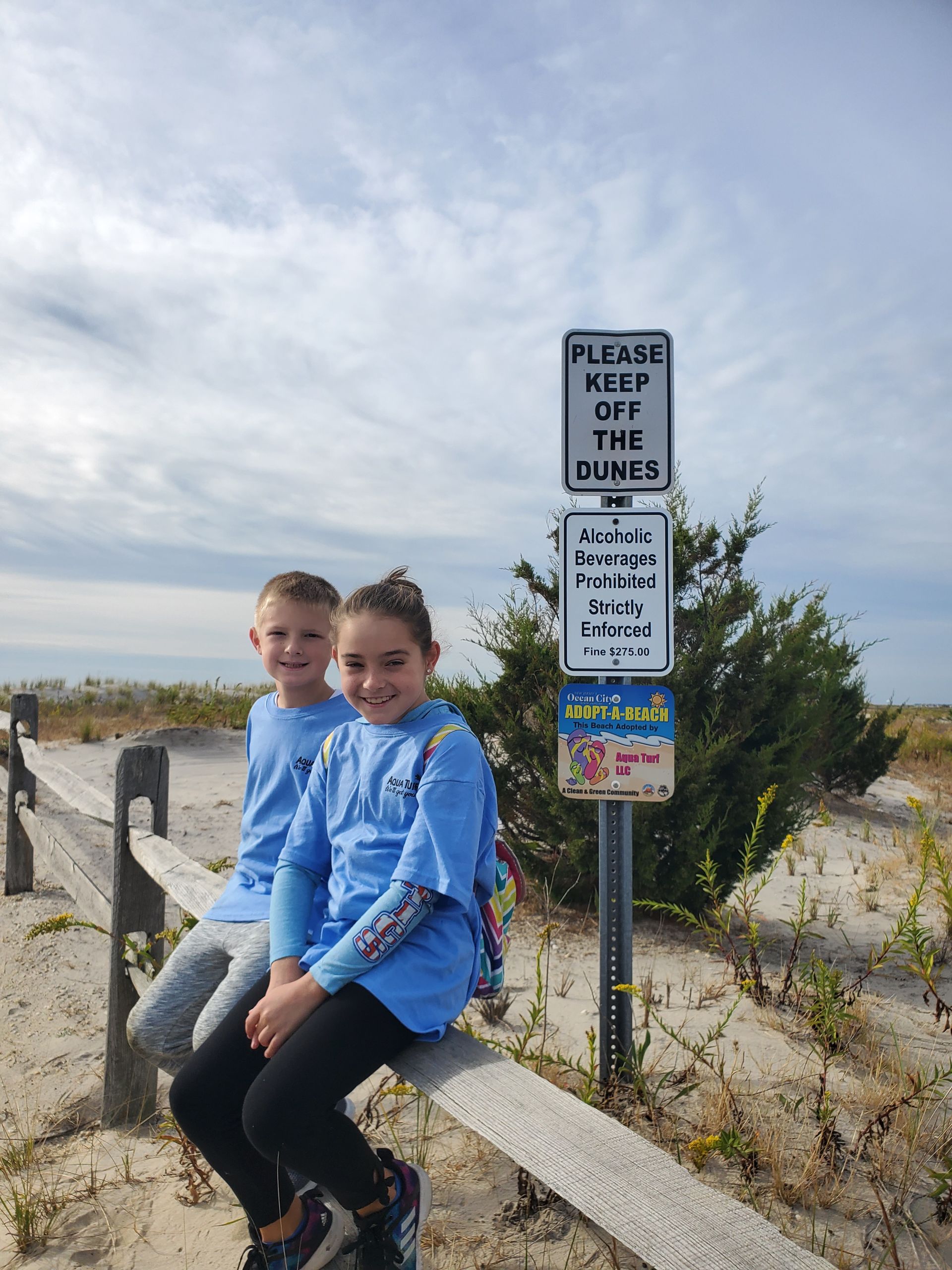 Two children in blue shirts sit on a wooden fence next to a sign that reads 