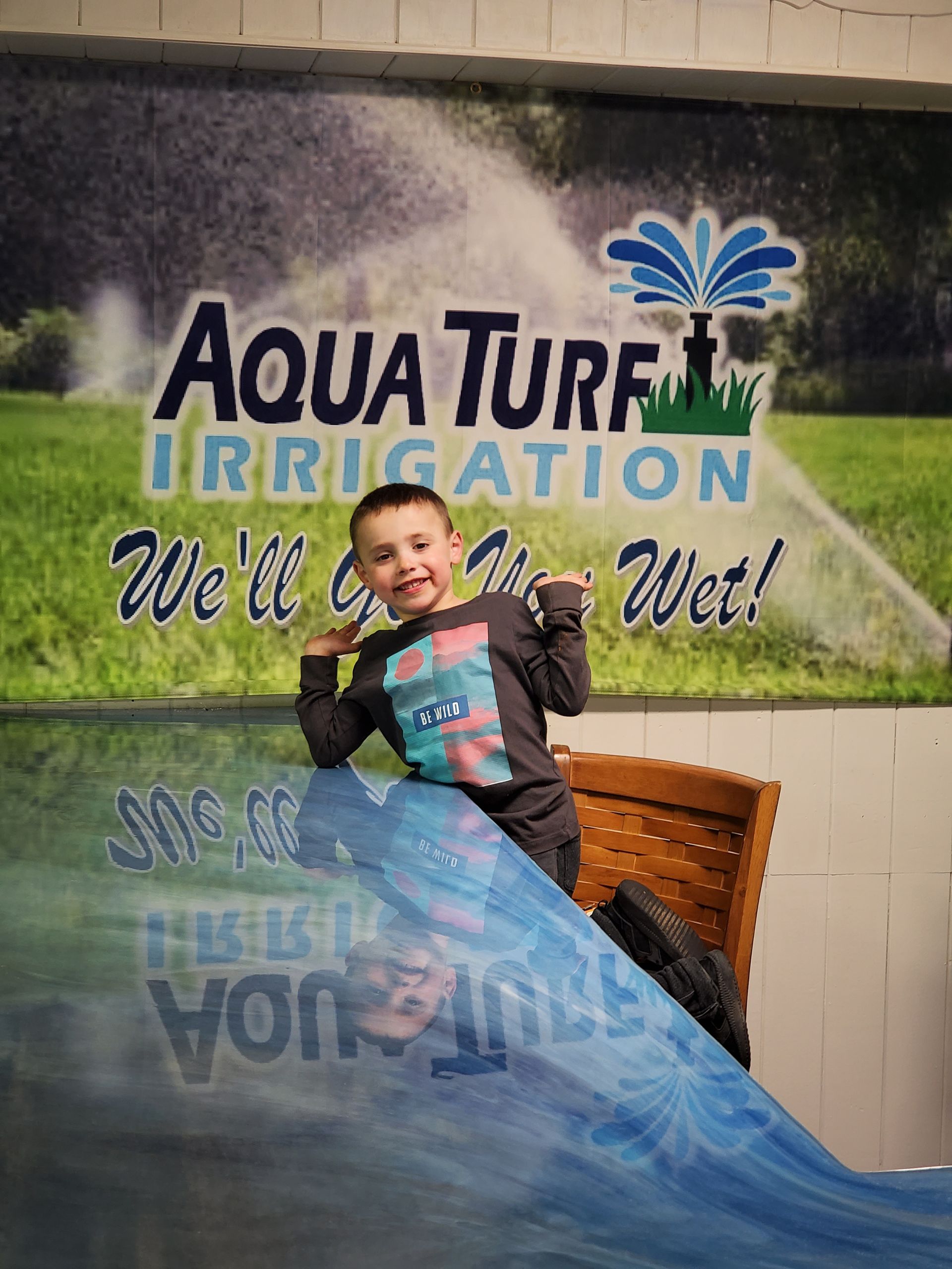 Young boy smiles, poses in front of Aqua Turf Irrigation sign. Blue and green background, water effect in foreground.