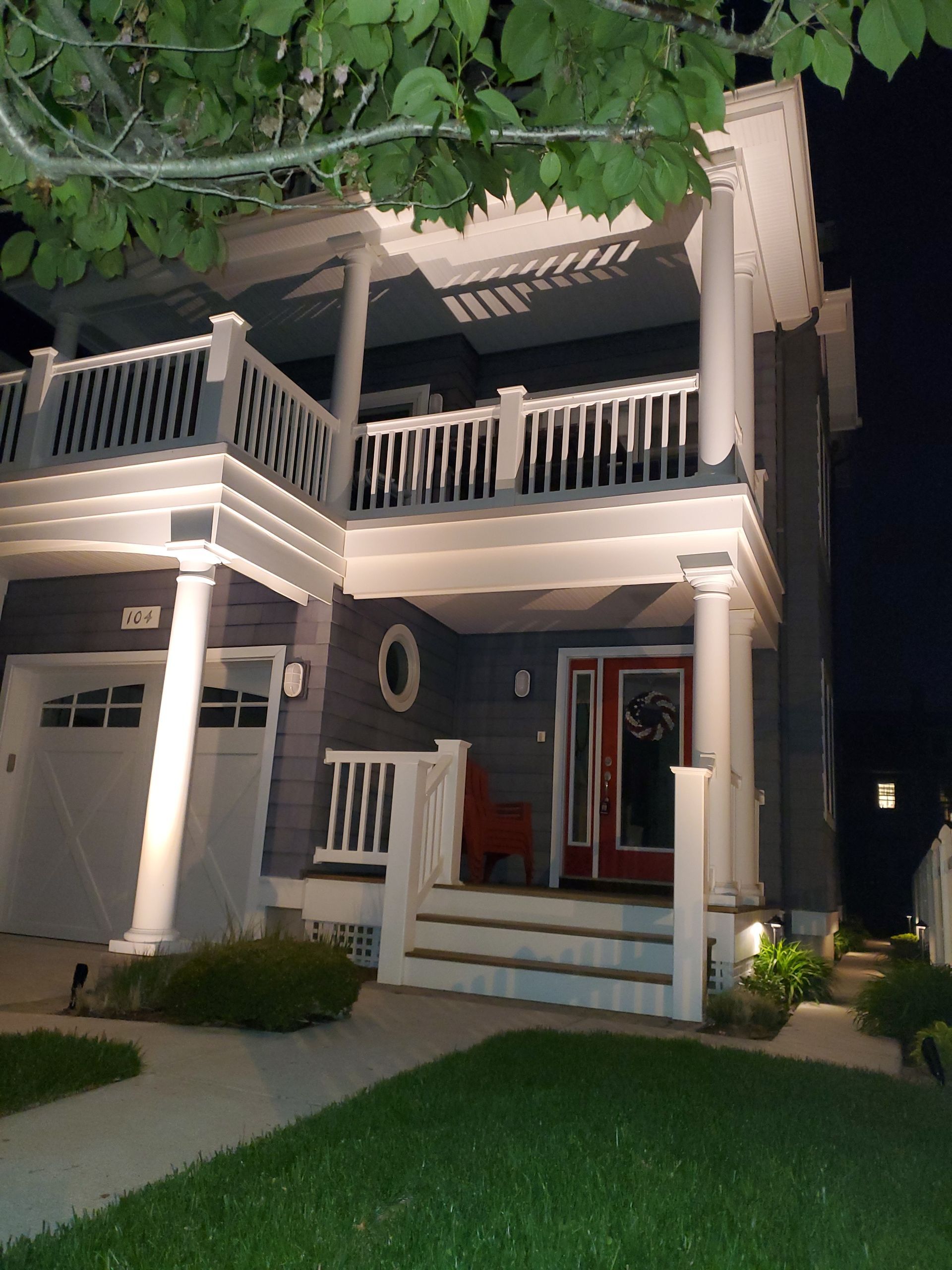 Two-story house with white trim, porch, and a red front door. Garage door on the left, sidewalk, and grass in front.