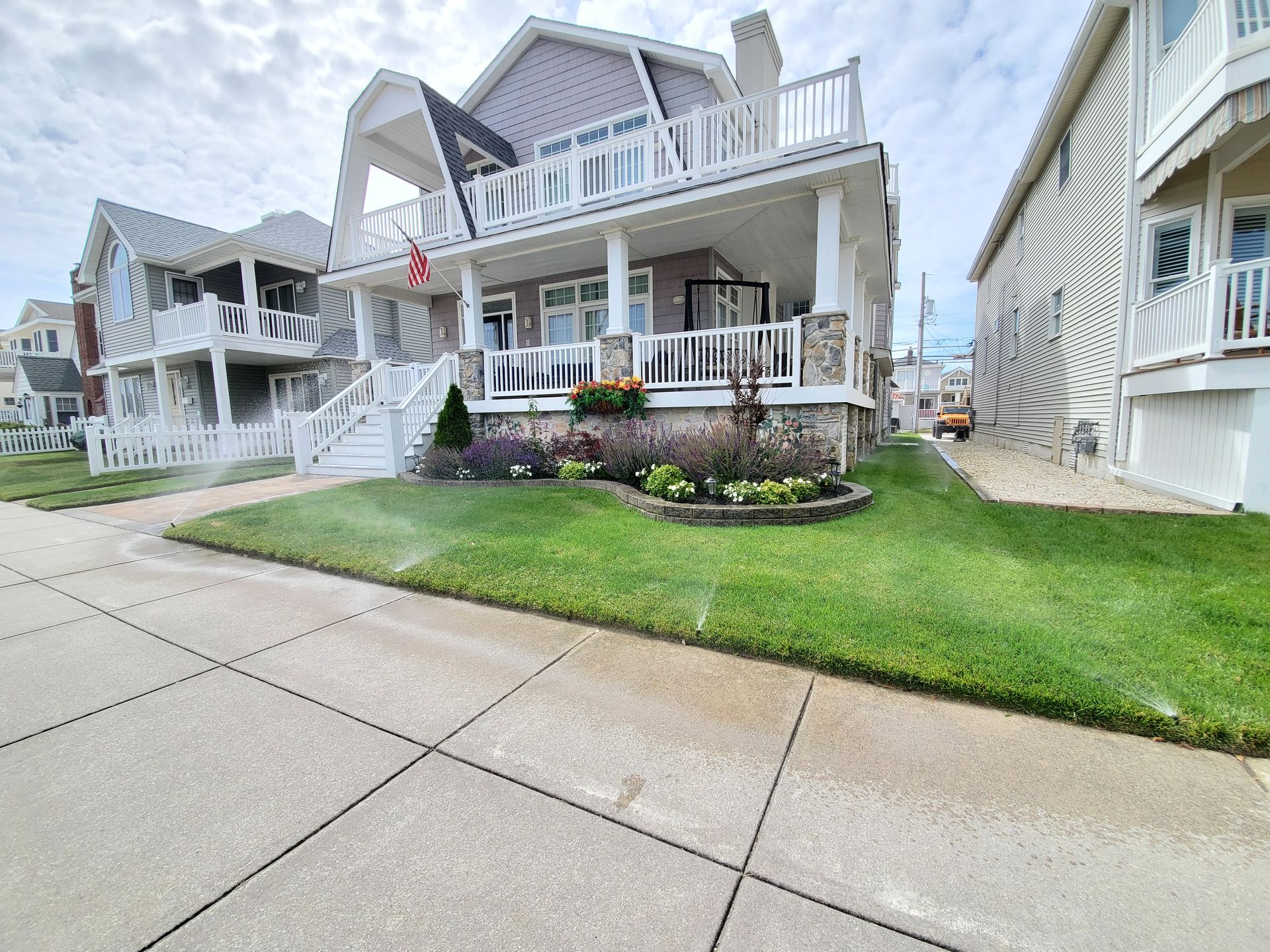 Two-story beach house with tan siding, porch, and grass lawn. Sprinklers water the green grass.