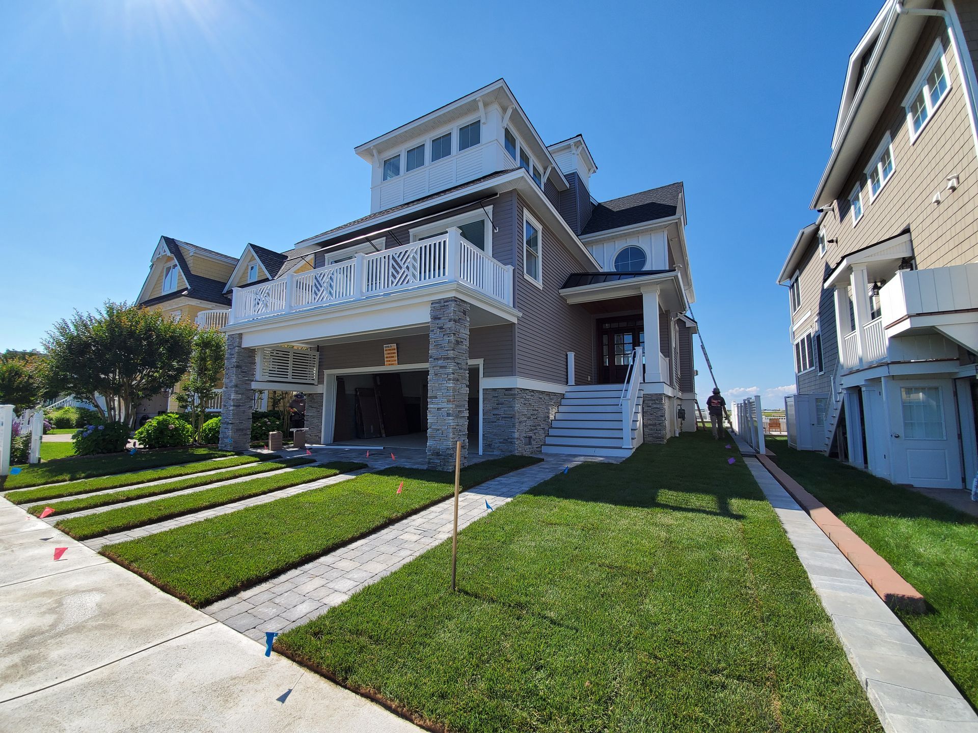 Multi-story house with gray siding, stone pillars, a balcony, and a manicured lawn under a blue sky.