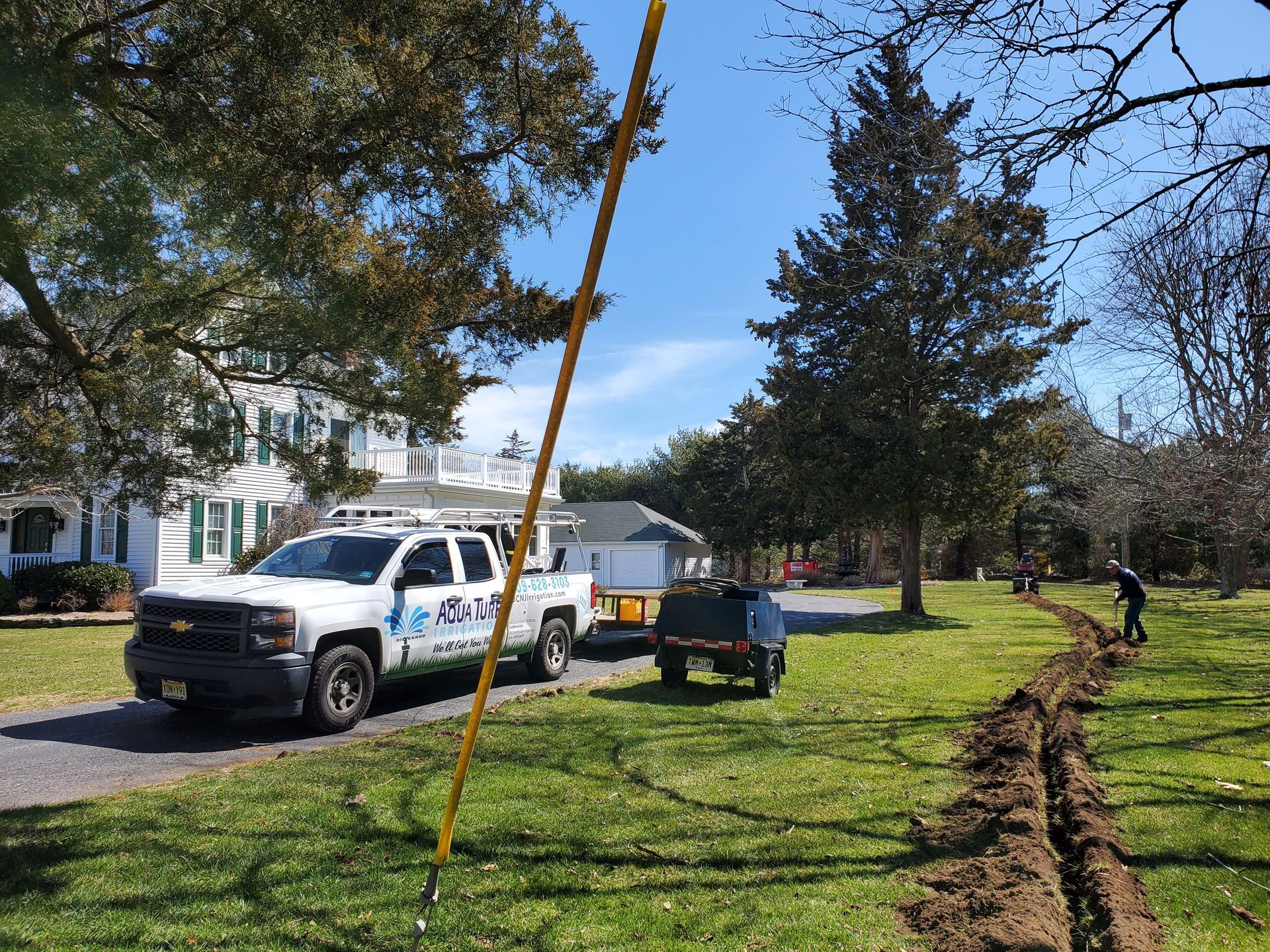 Utility truck and trailer parked on grass, next to a trench being dug in a yard near a house.
