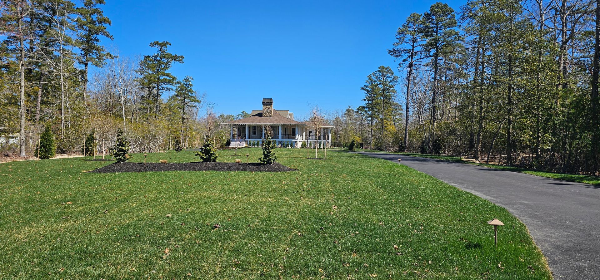 A long driveway leads to a white house with a porch, framed by trees and green lawn under a blue sky.