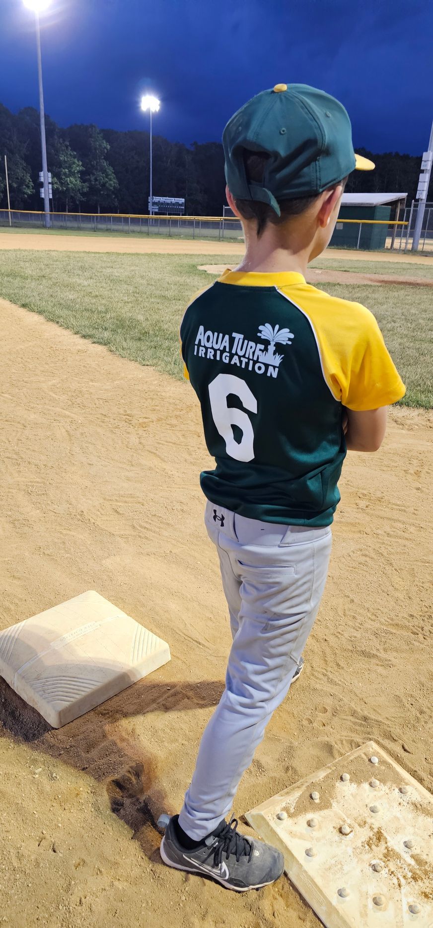 A child in a baseball uniform stands on the field, facing away, arms crossed. The number 6 is visible.