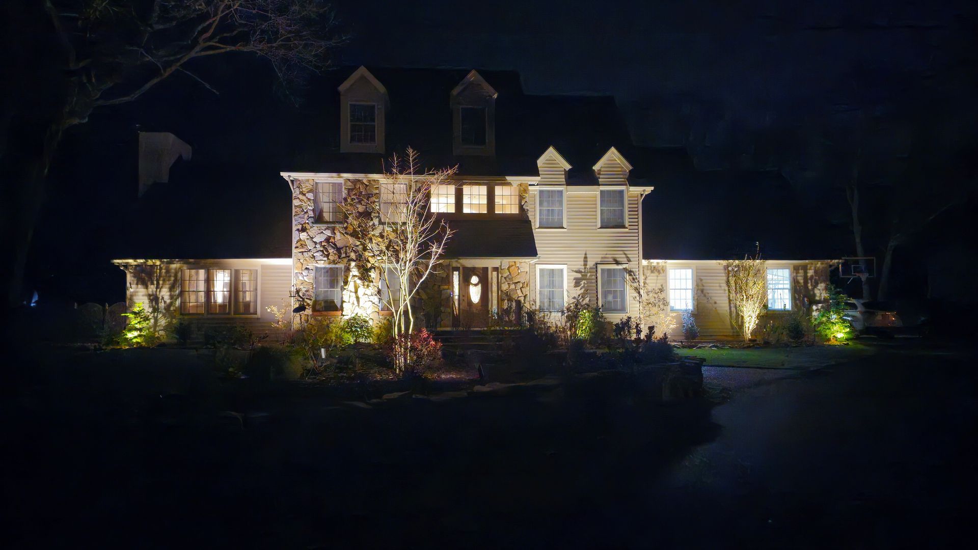 Large two-story house at night with lights on, shrubs and driveway in front.