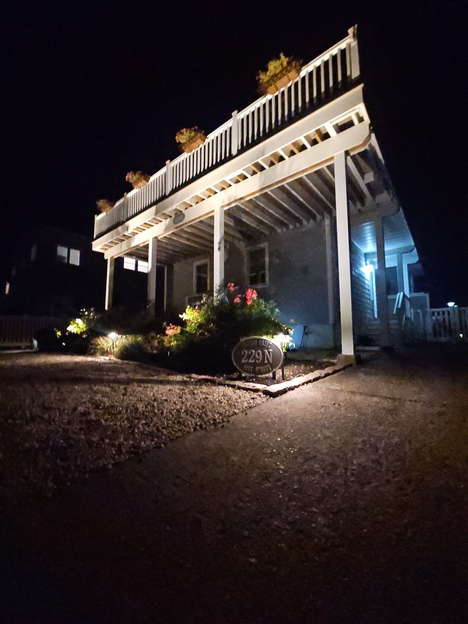 House at night, lit from below with a second-story deck, flowers and shrubs in front.