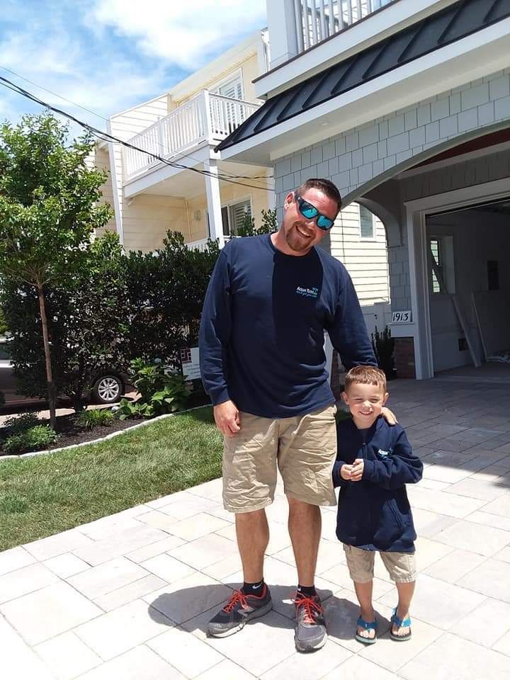 Man and child stand on a paved area in front of a house on a sunny day.