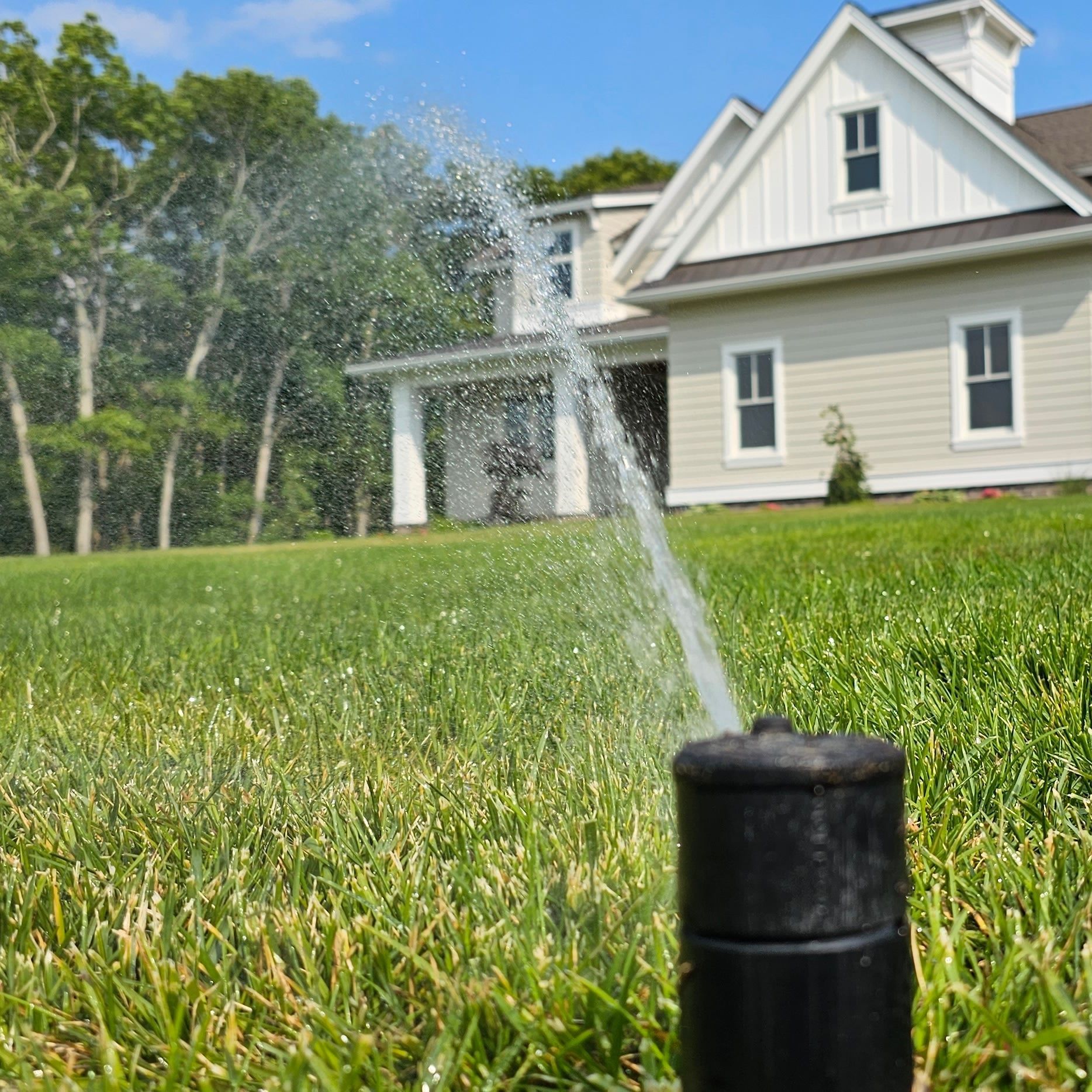 Sprinkler spraying water onto a green lawn in front of a beige house with white trim.