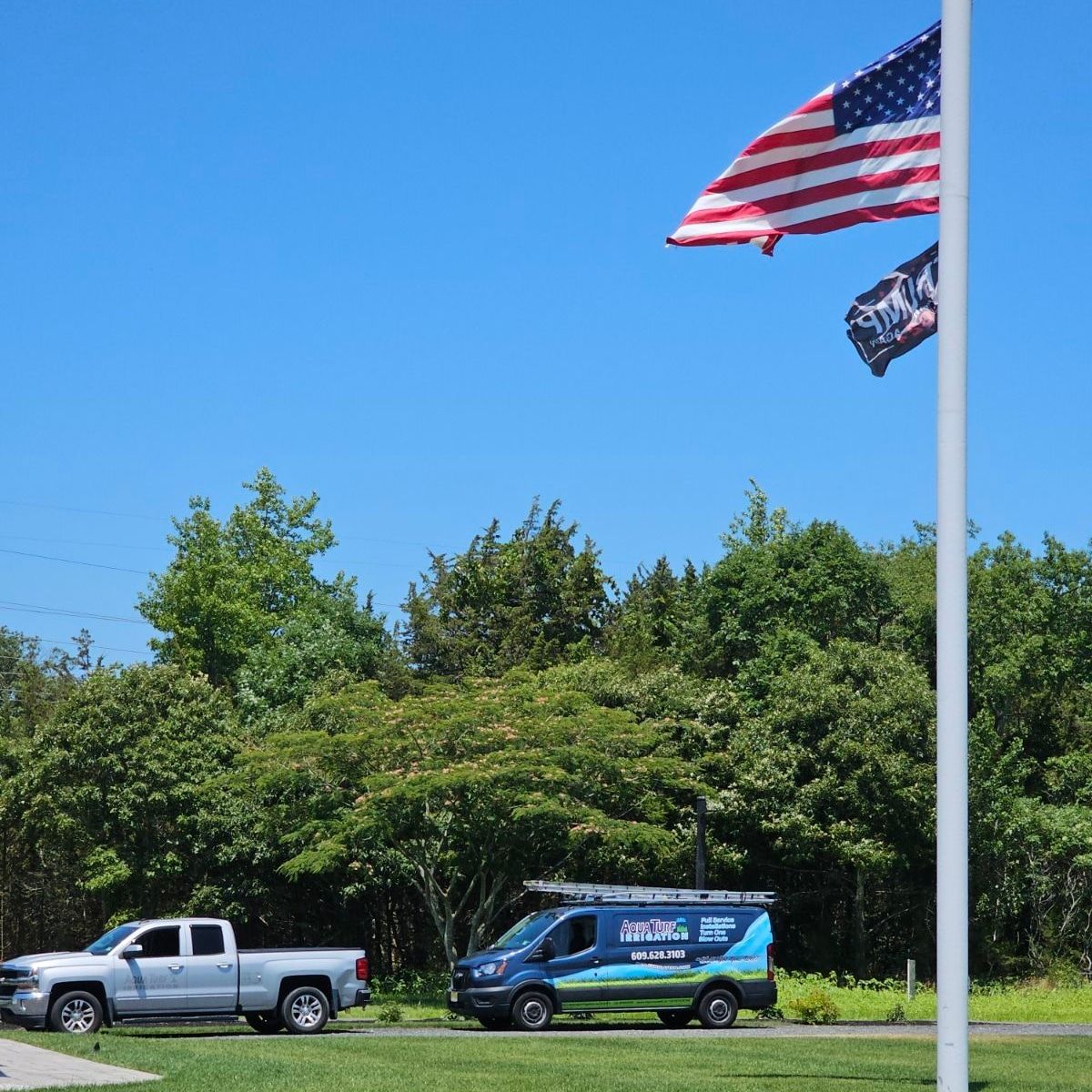 A silver pickup truck and blue van parked on grass under an American flag.