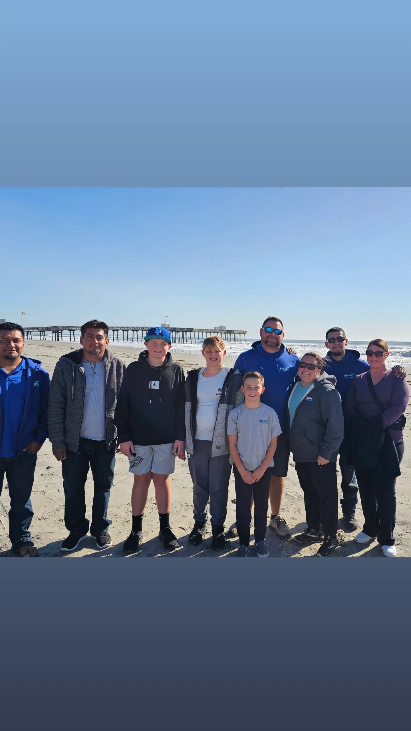 Group of people standing on a sandy beach in front of a pier on a sunny day.