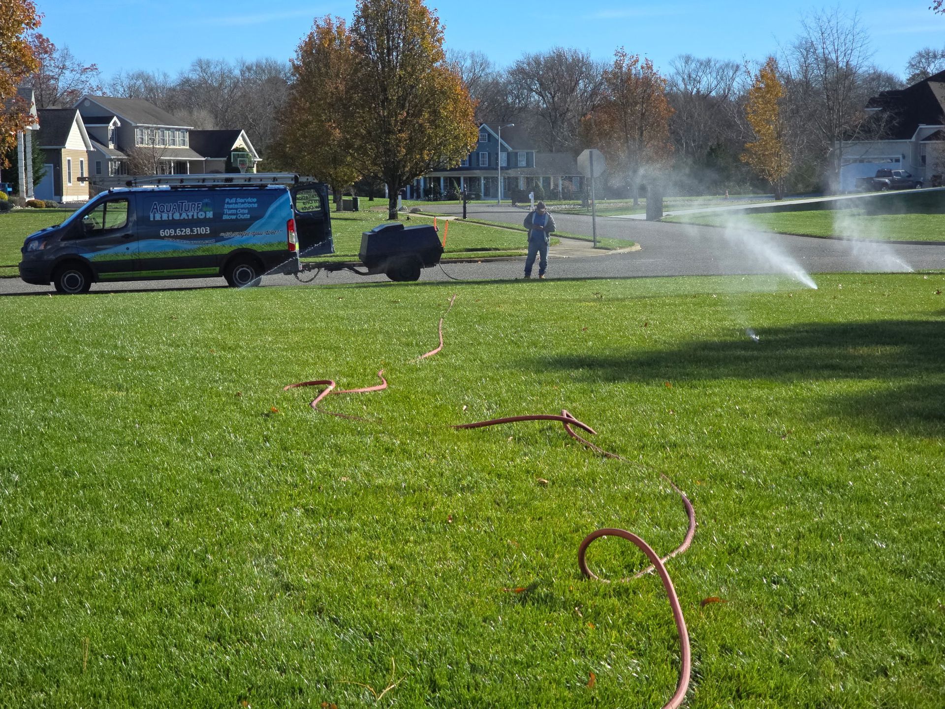 Sprinkler system repair: Van, man, and water spraying on a green lawn in a suburban neighborhood.