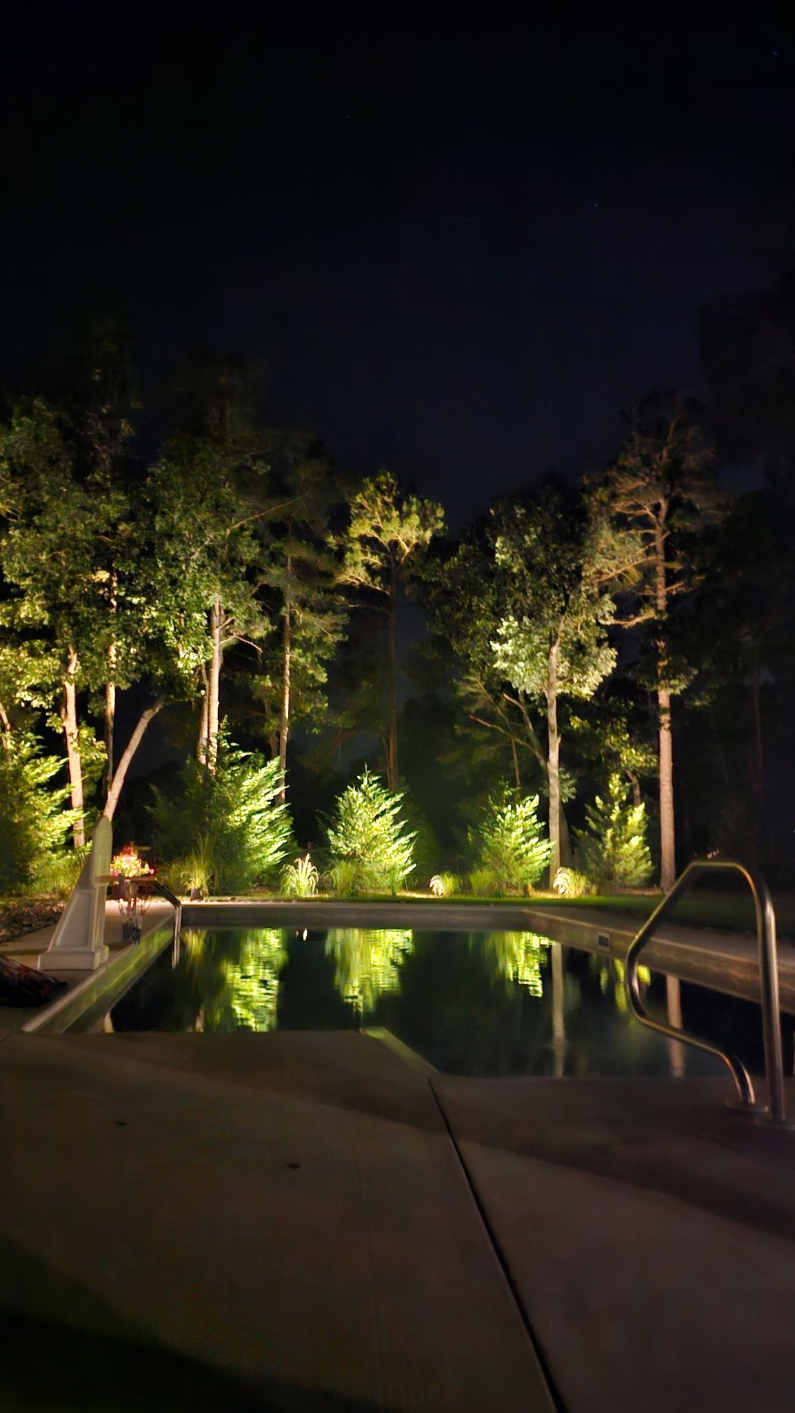 Nighttime view of a pool with landscape lighting illuminating trees in a wooded area. The pool water reflects the lights.