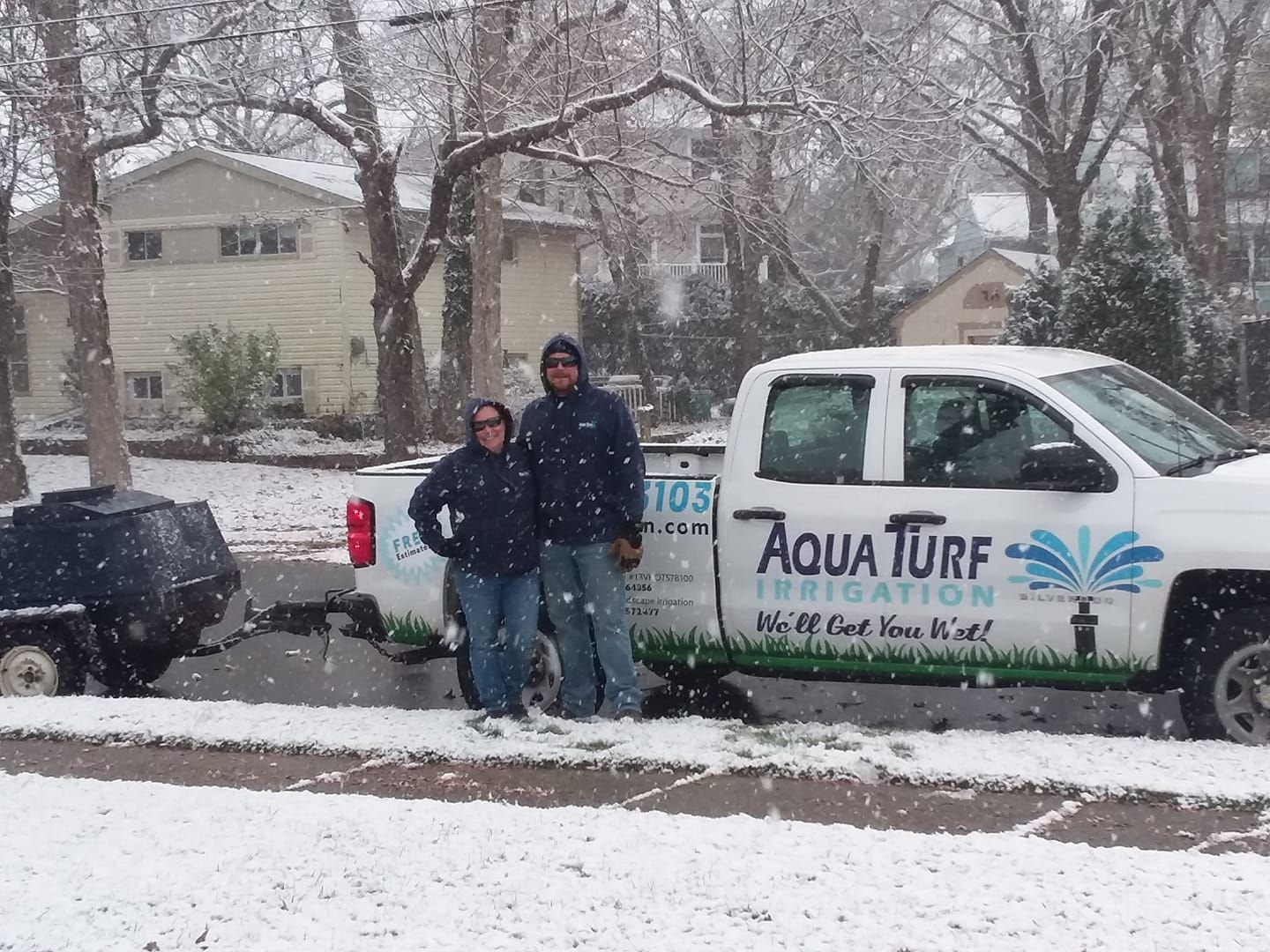 Two people stand beside a white Aqua Turf Irrigation truck in a snowy residential setting.