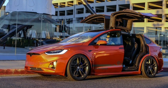 an orange Tesla Model X with its doors open is parked on the side of the road.