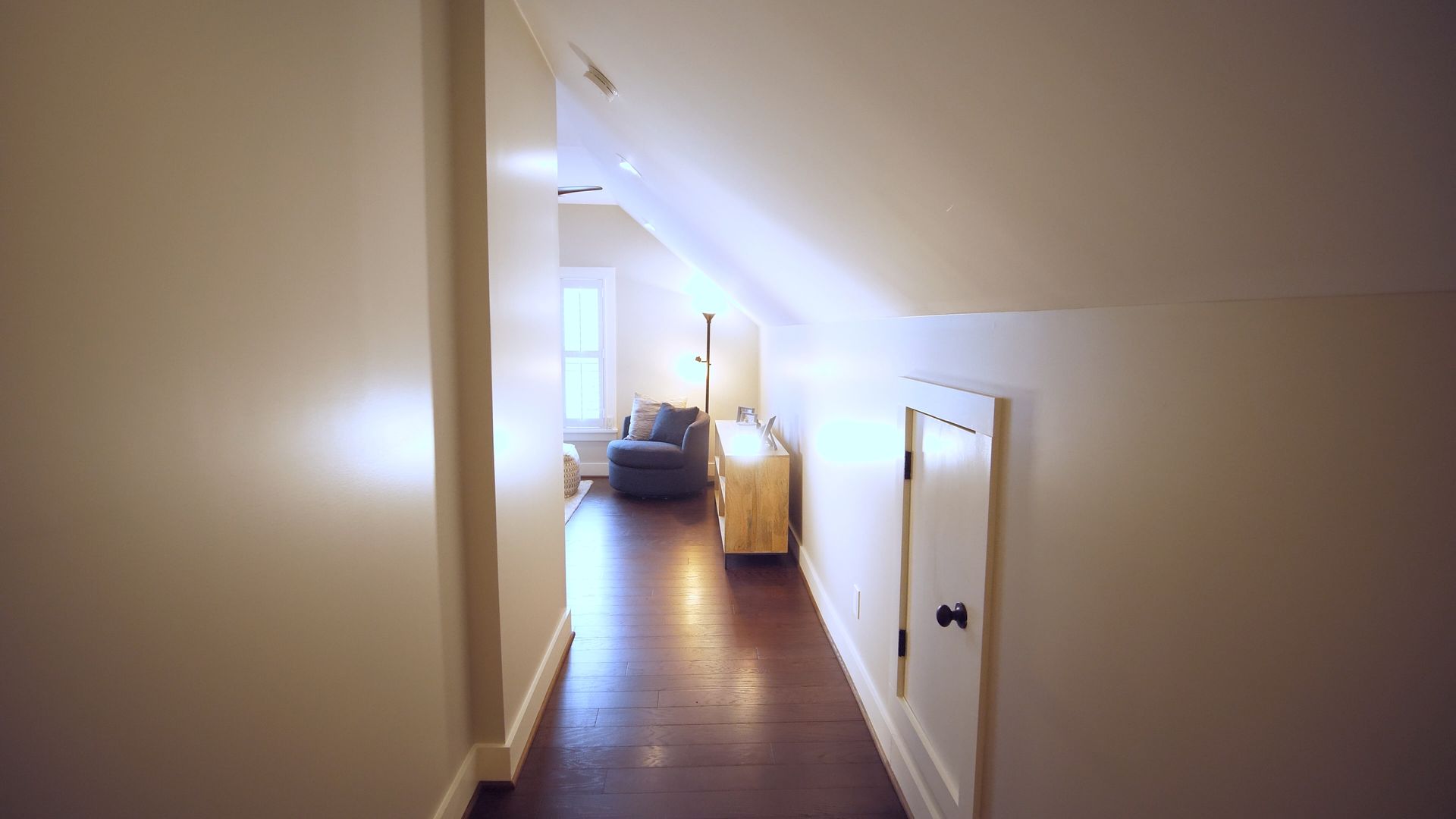 Narrow hallway with angled ceilings, leading to a room with a window, hardwood floor, and a small storage door.