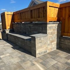 A patio with gray pavers, a brick-style retaining wall, and a wooden fence in sunlight.