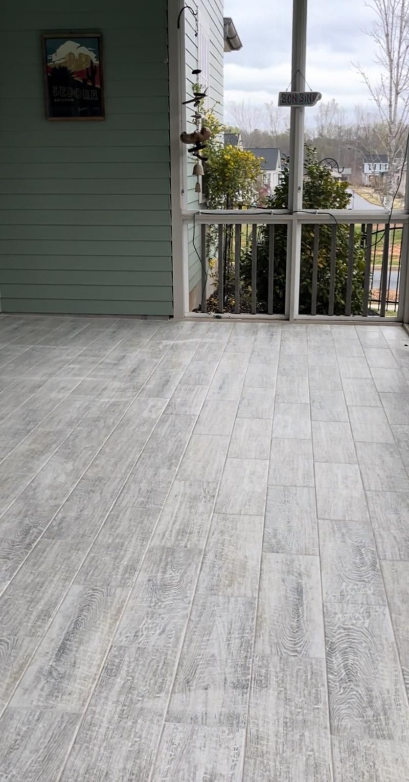 A covered porch with gray wood-look tile flooring. Light blue siding, white railing, and a landscape view.