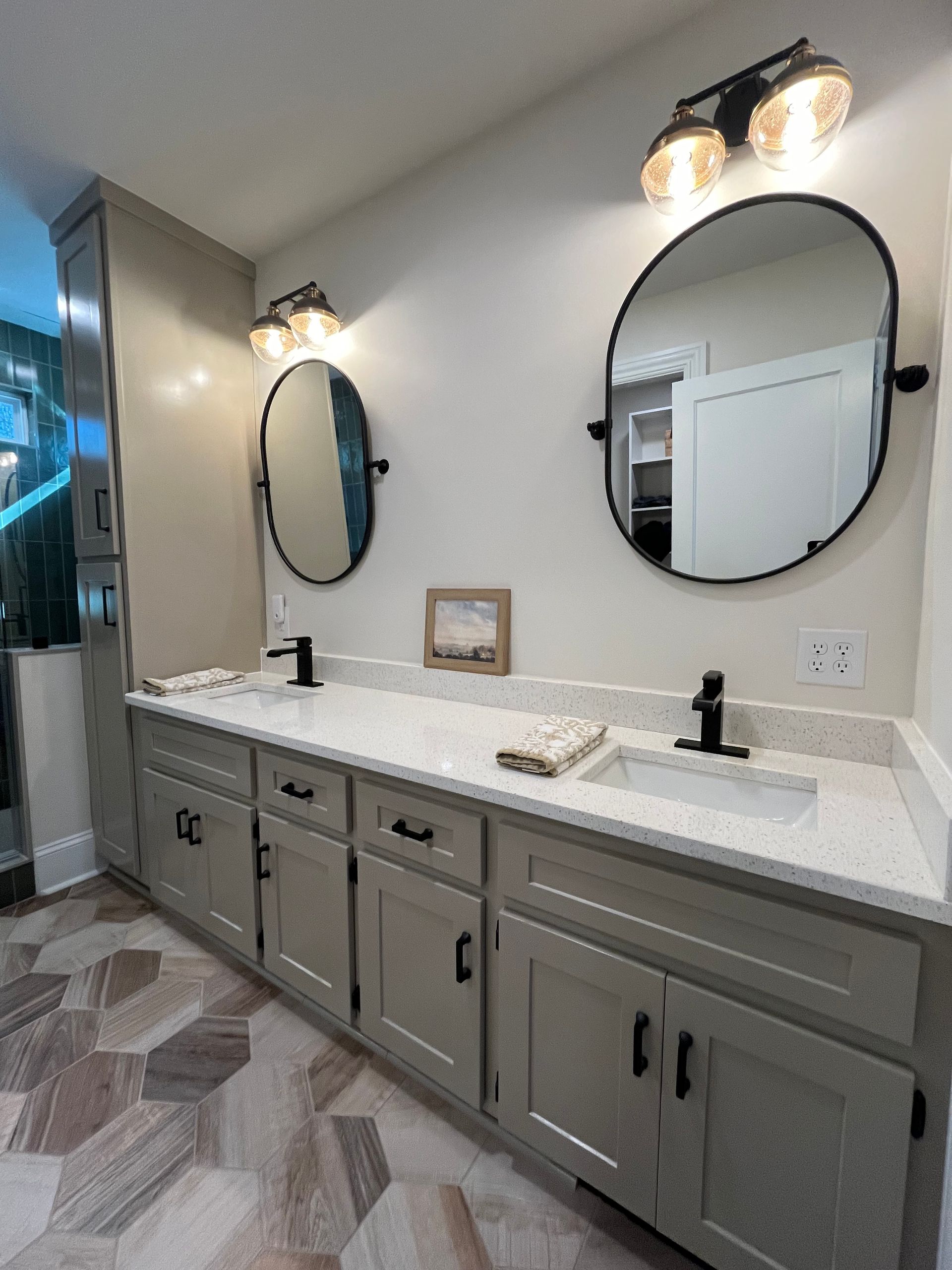 Bathroom with light gray cabinets, oval mirrors, black fixtures, and patterned tile flooring.