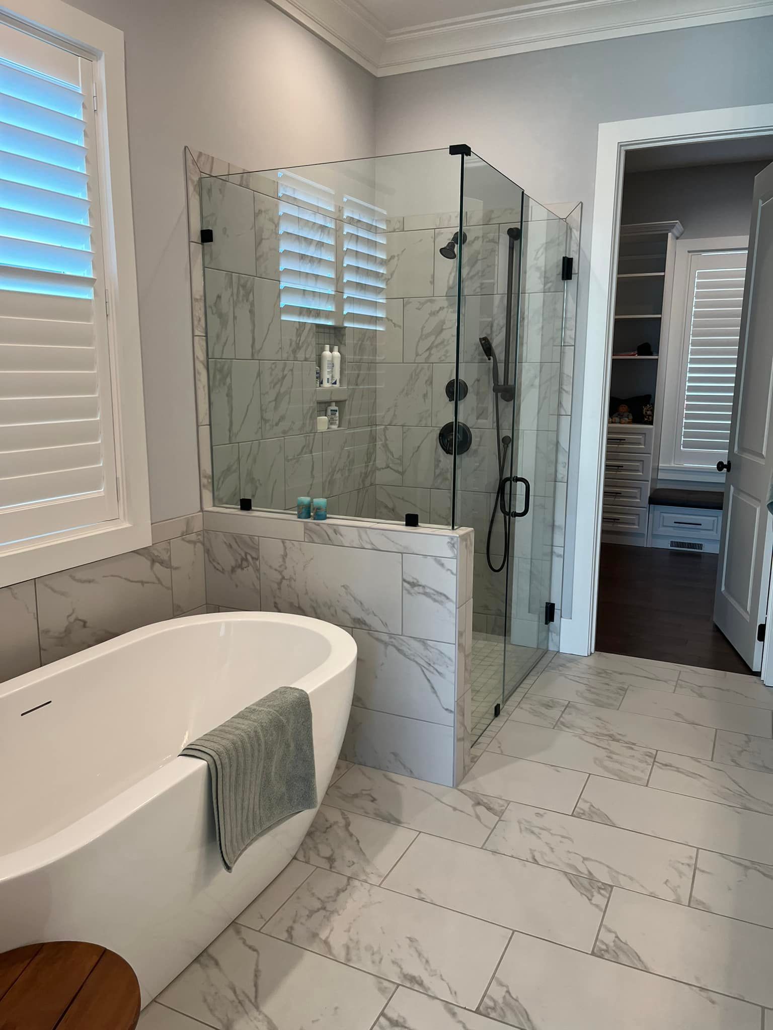 Modern bathroom: white soaking tub, marble tile shower with black fixtures, window with shutters.