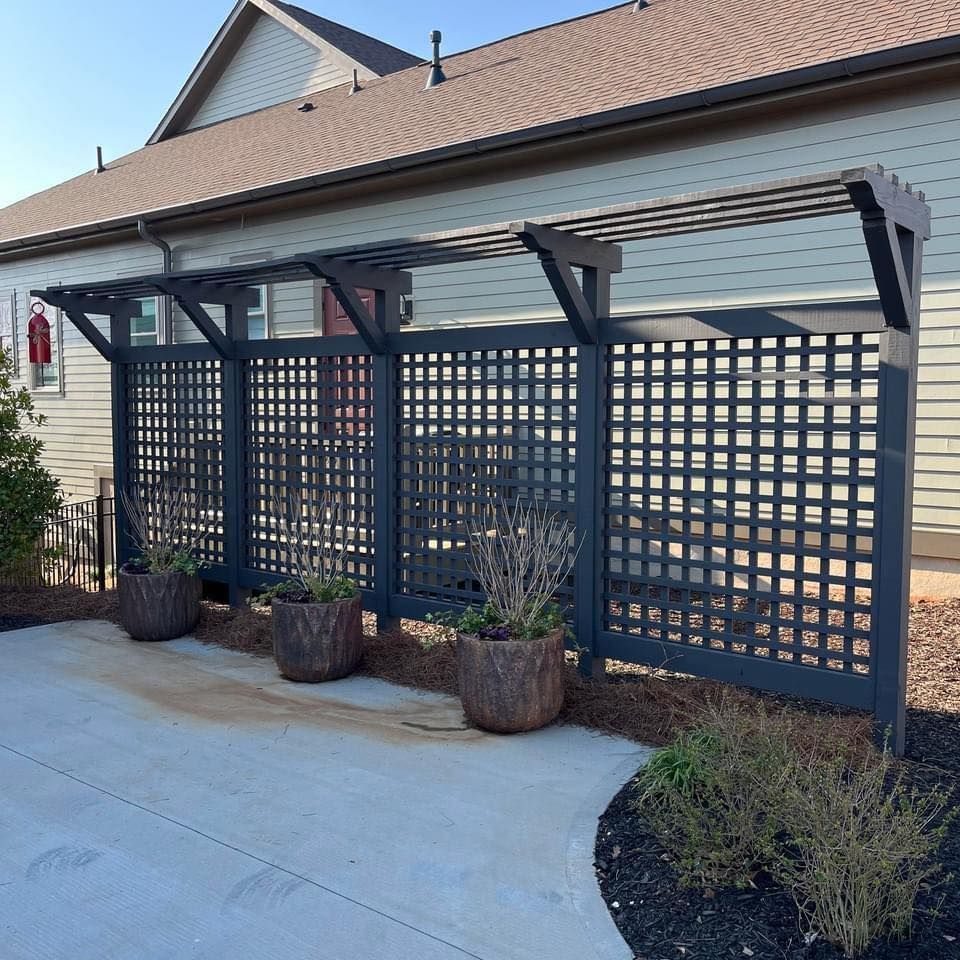 Black lattice privacy fence with overhead pergola, planters along gray paved area.