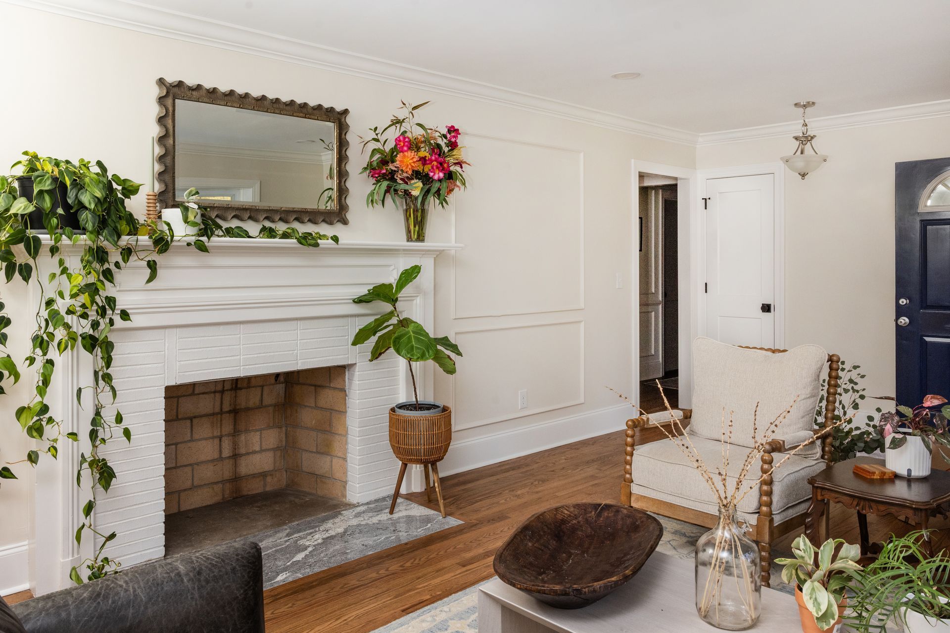 Living room with fireplace, mirror, plants, white walls, wood floor, and blue door.