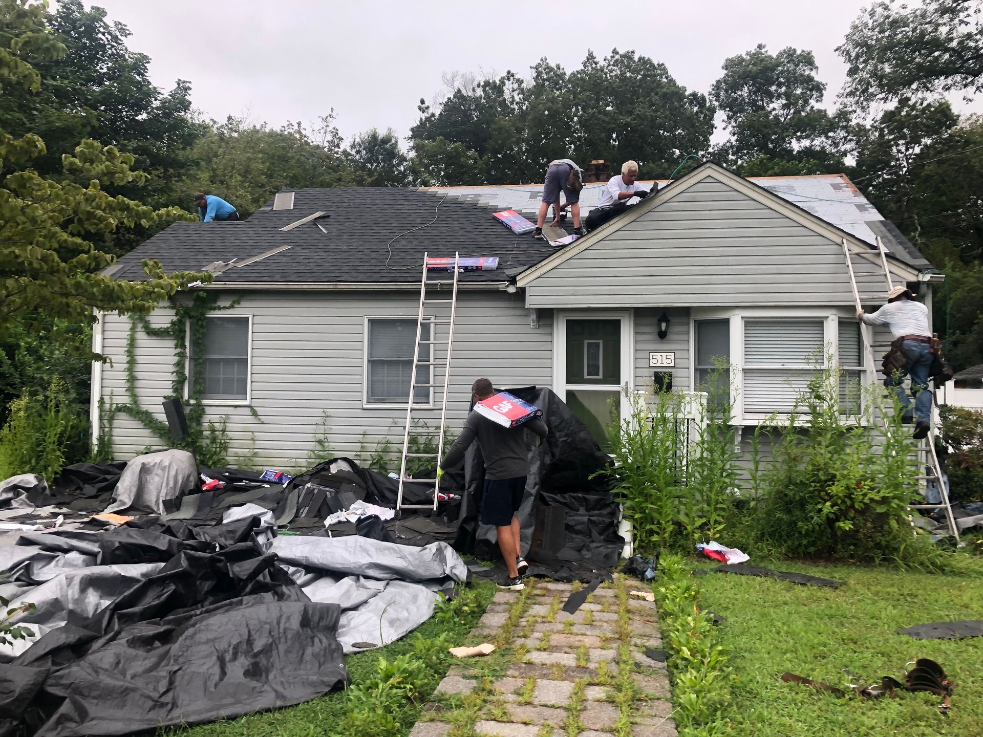 People repairing a gray house roof; ladders, tarp, and debris on the lawn.
