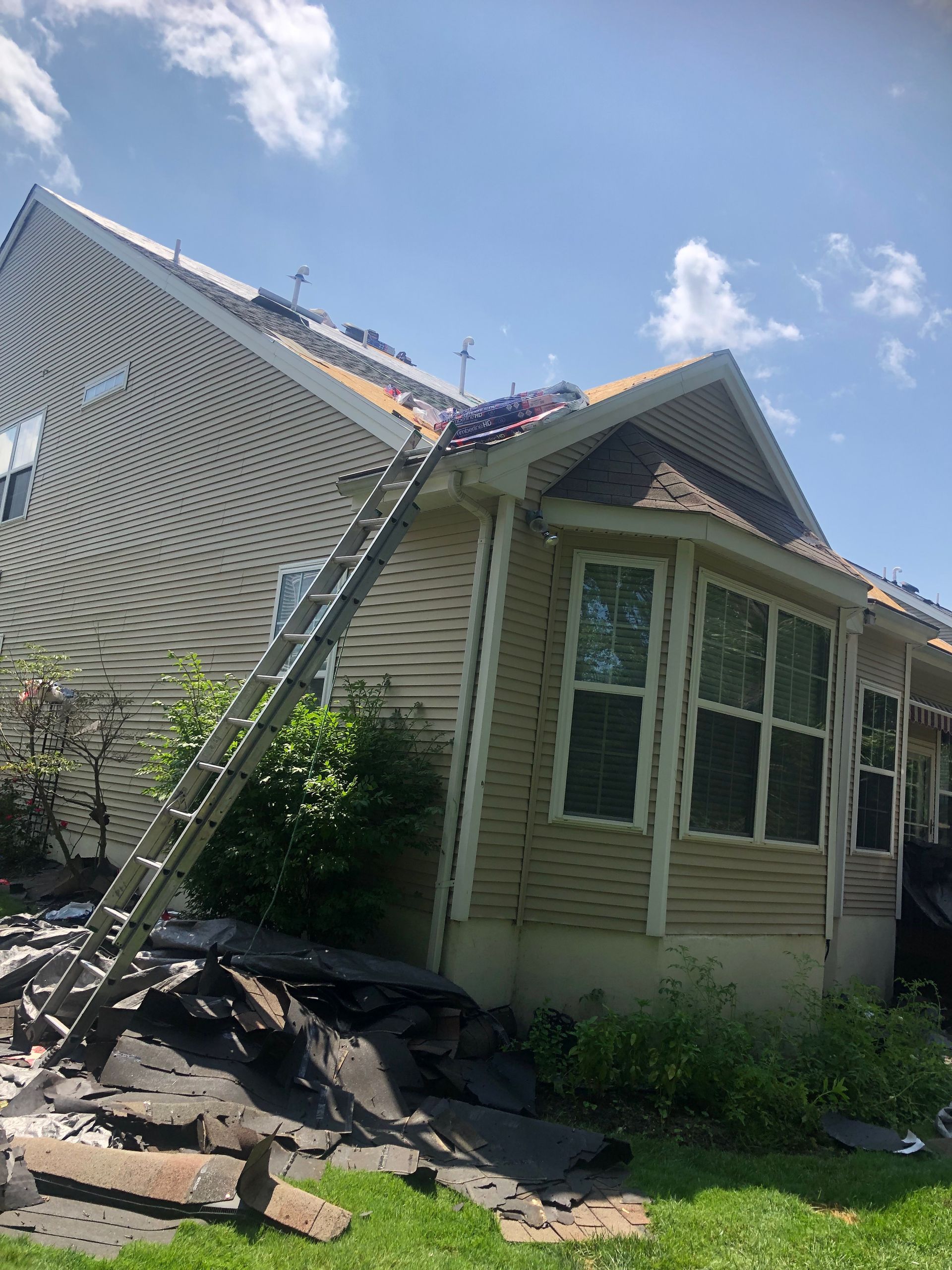 A ladder leans against a house with a partially replaced roof. Roofing debris is on the ground.