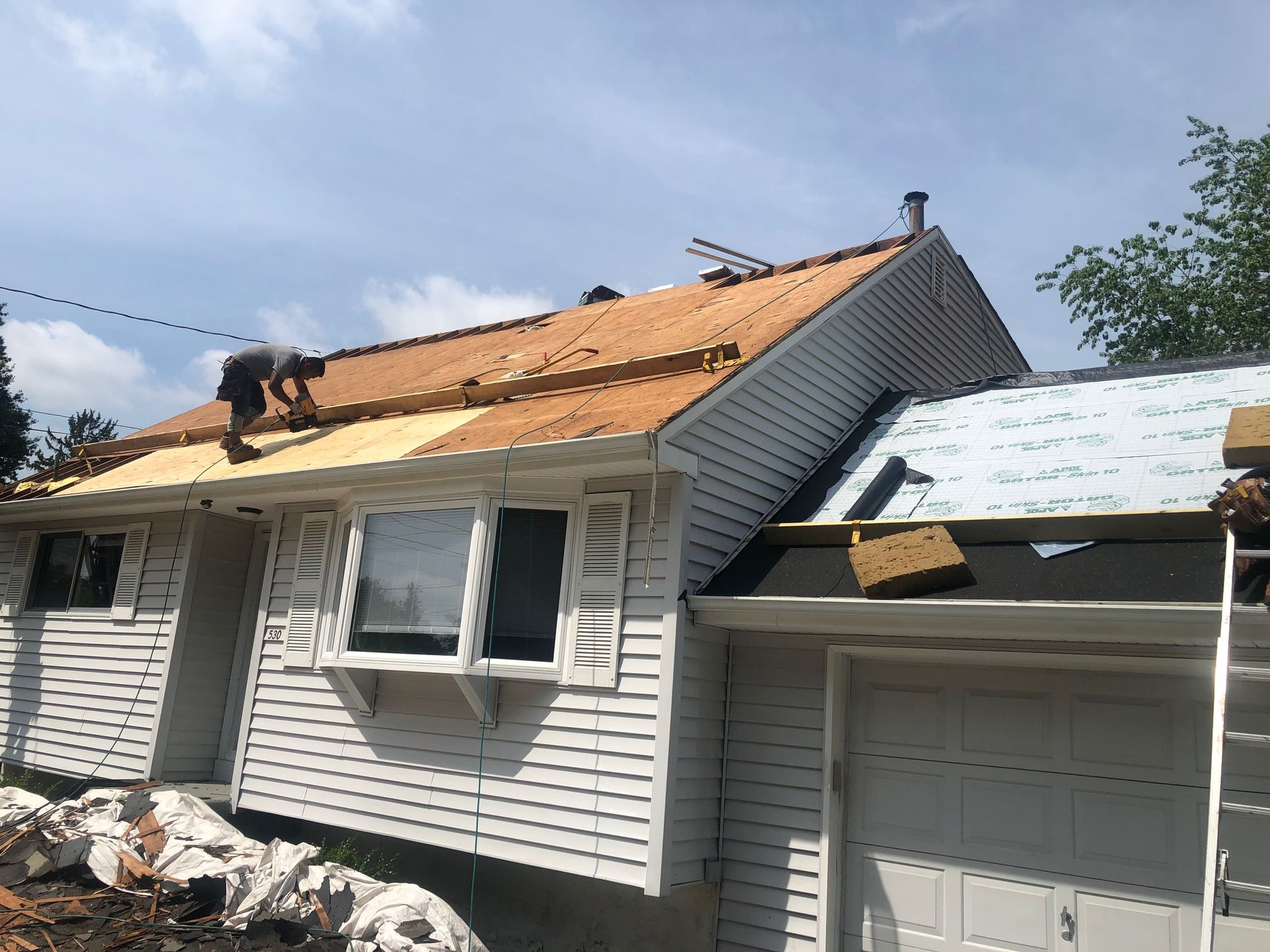 Construction worker replacing roof shingles on a house with white siding under a blue sky.