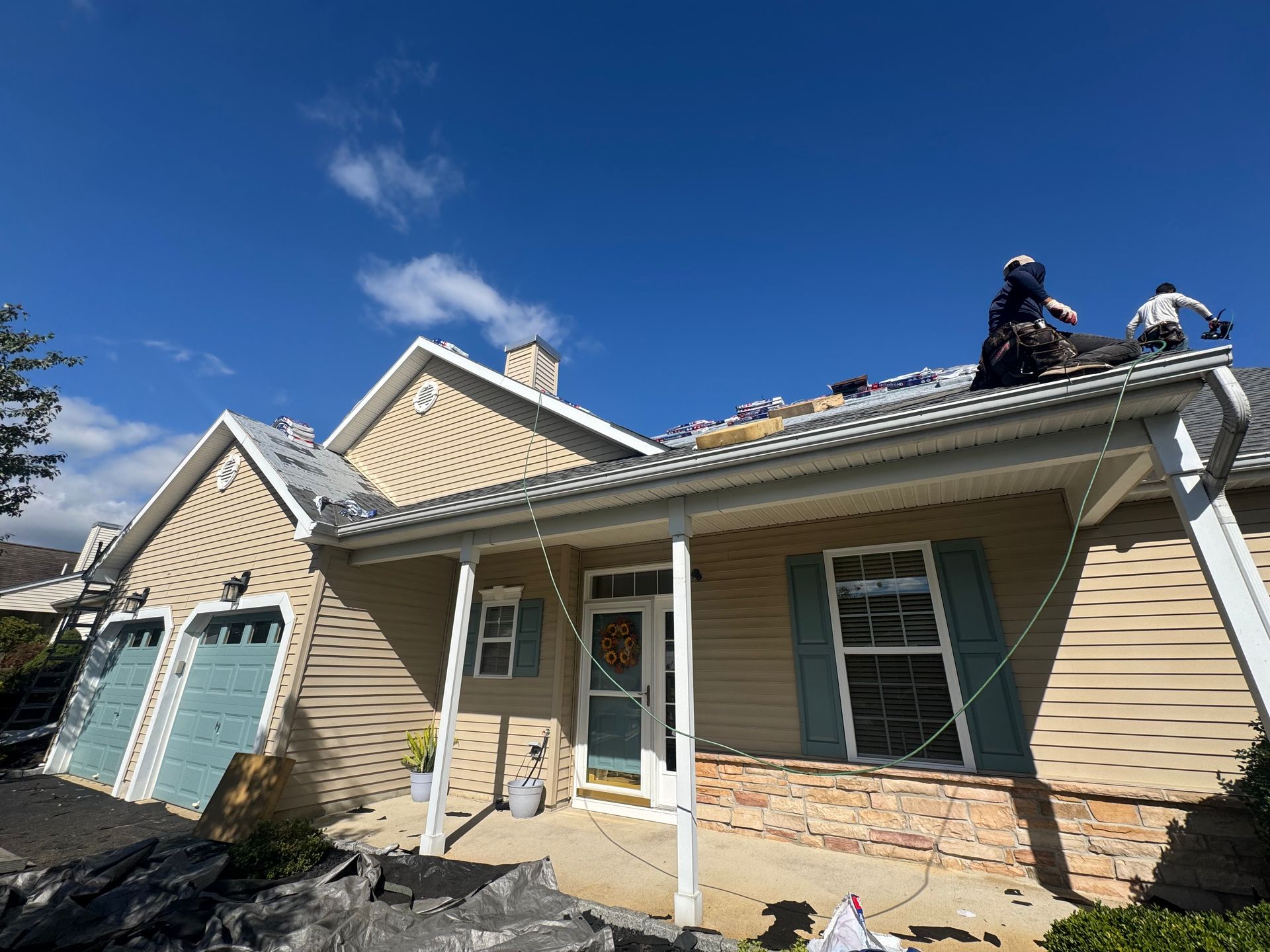 Roofers working on a house with beige siding and blue garage doors under a bright blue sky.