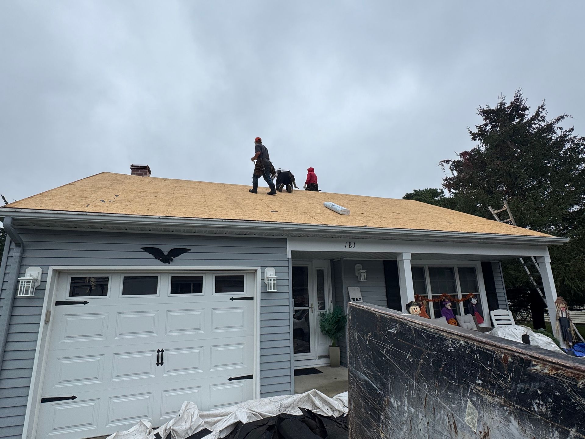 Three people on a roof removing shingles. Gray house with white garage door, cloudy sky.