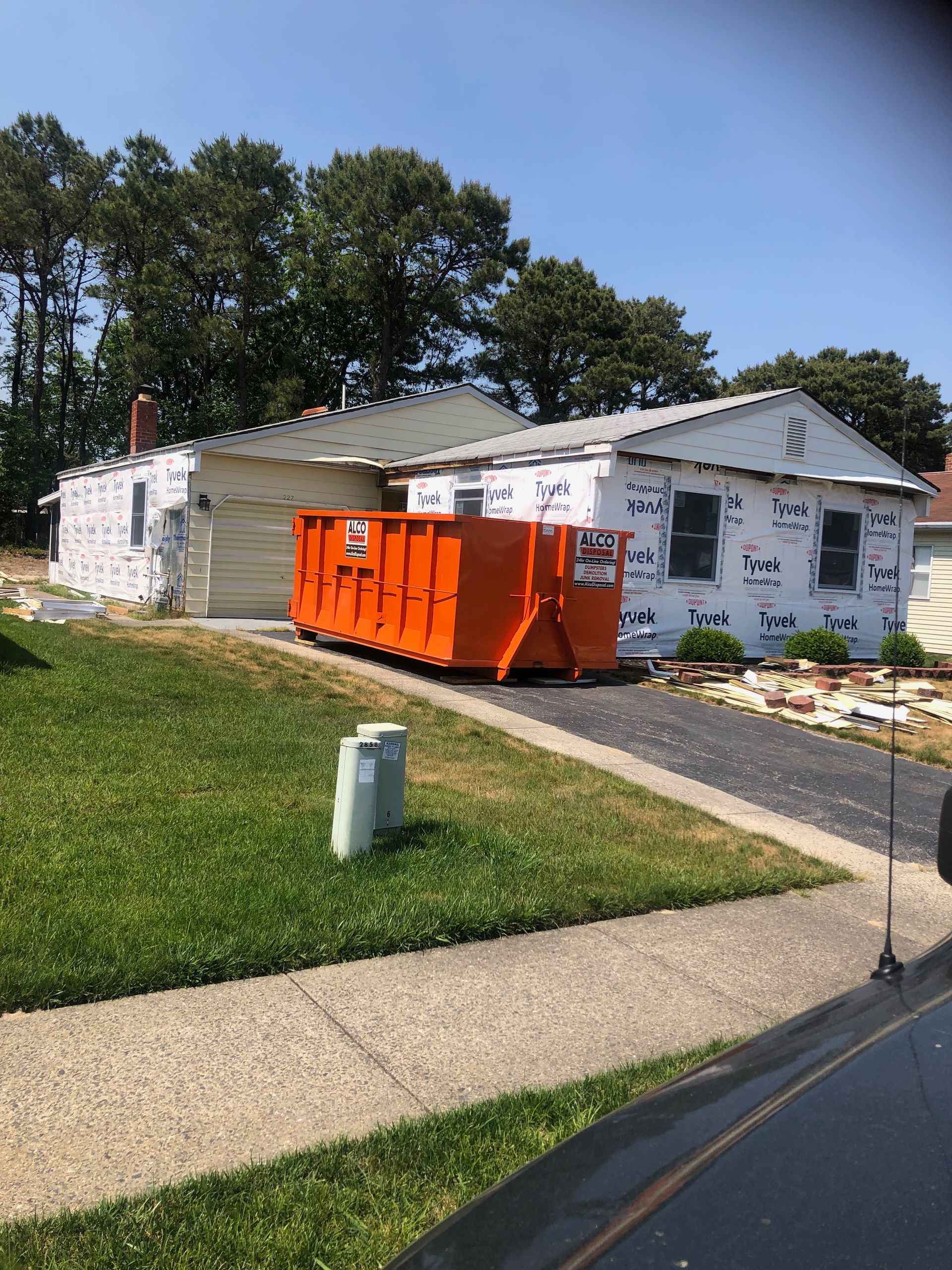 House under construction, orange dumpster in driveway, covered with wrap; green grass, clear sky.