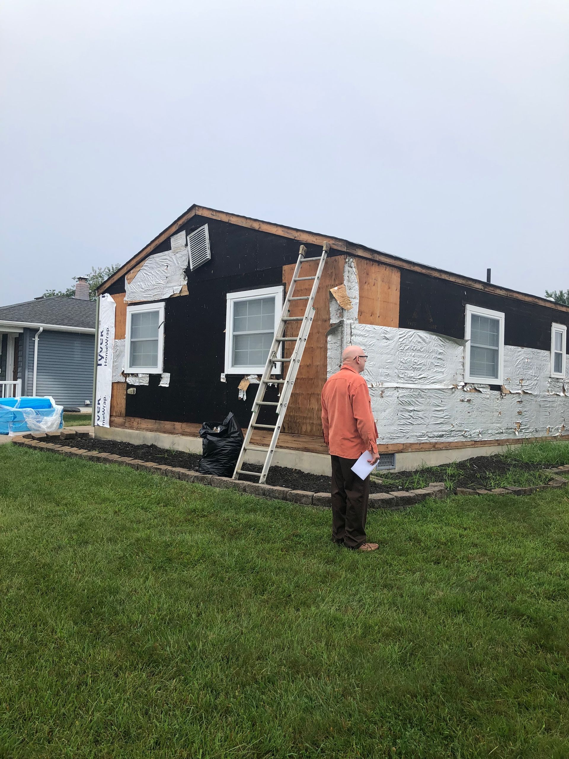 House under renovation; siding partially removed. A man stands outside, ladder propped against the building, overcast sky.