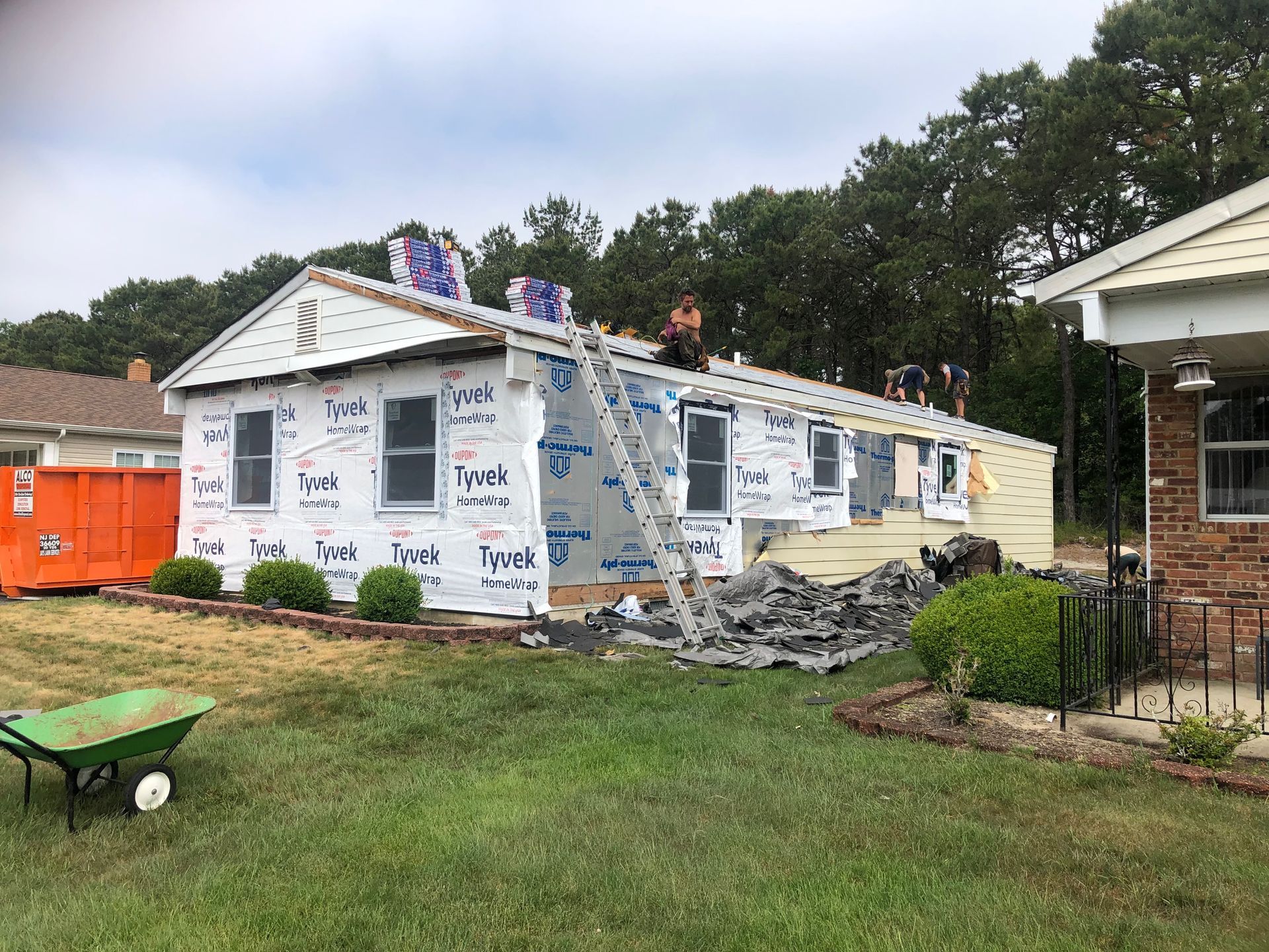 House under construction with workers, exposed framing, and protective wrap, surrounded by debris and yard.