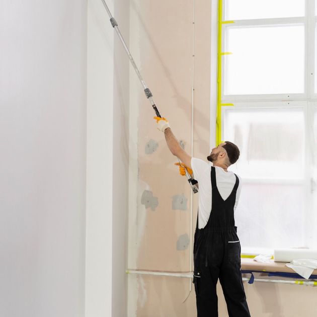 Person on a ladder painting a tall wall with a roller in a bright room