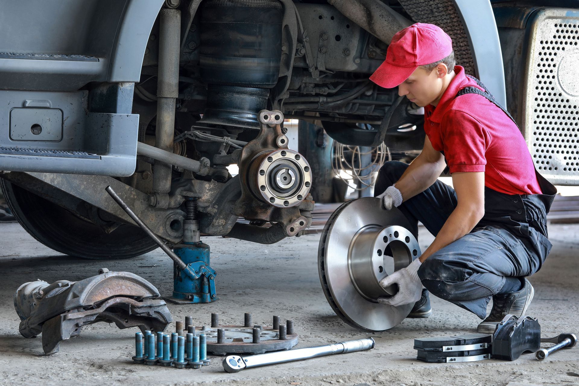 A mechanic in a red shirt and cap kneeling in a garage to inspect a truck's brake rotor.