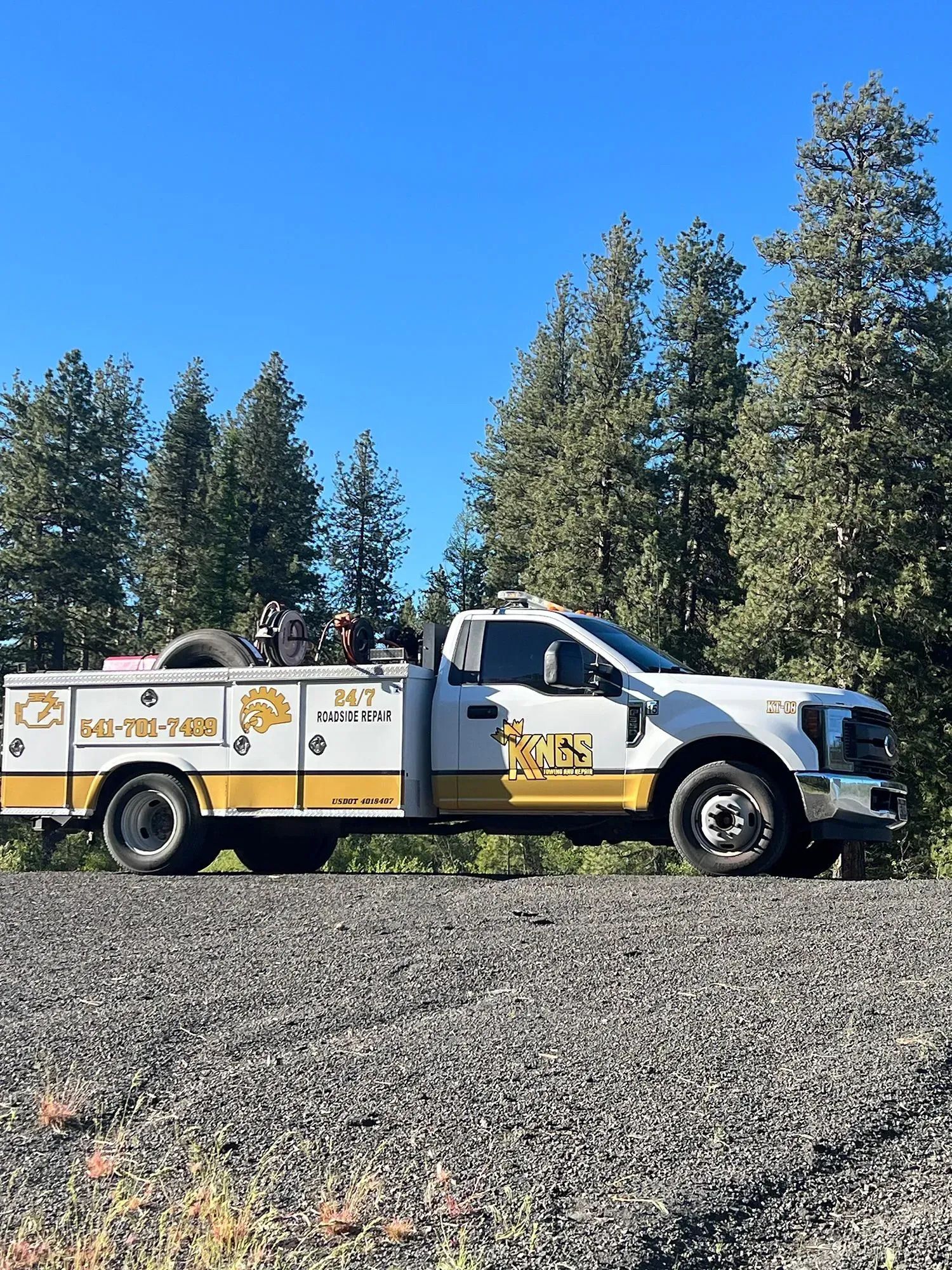 White utility truck parked on gravel in front of evergreen trees under a blue sky