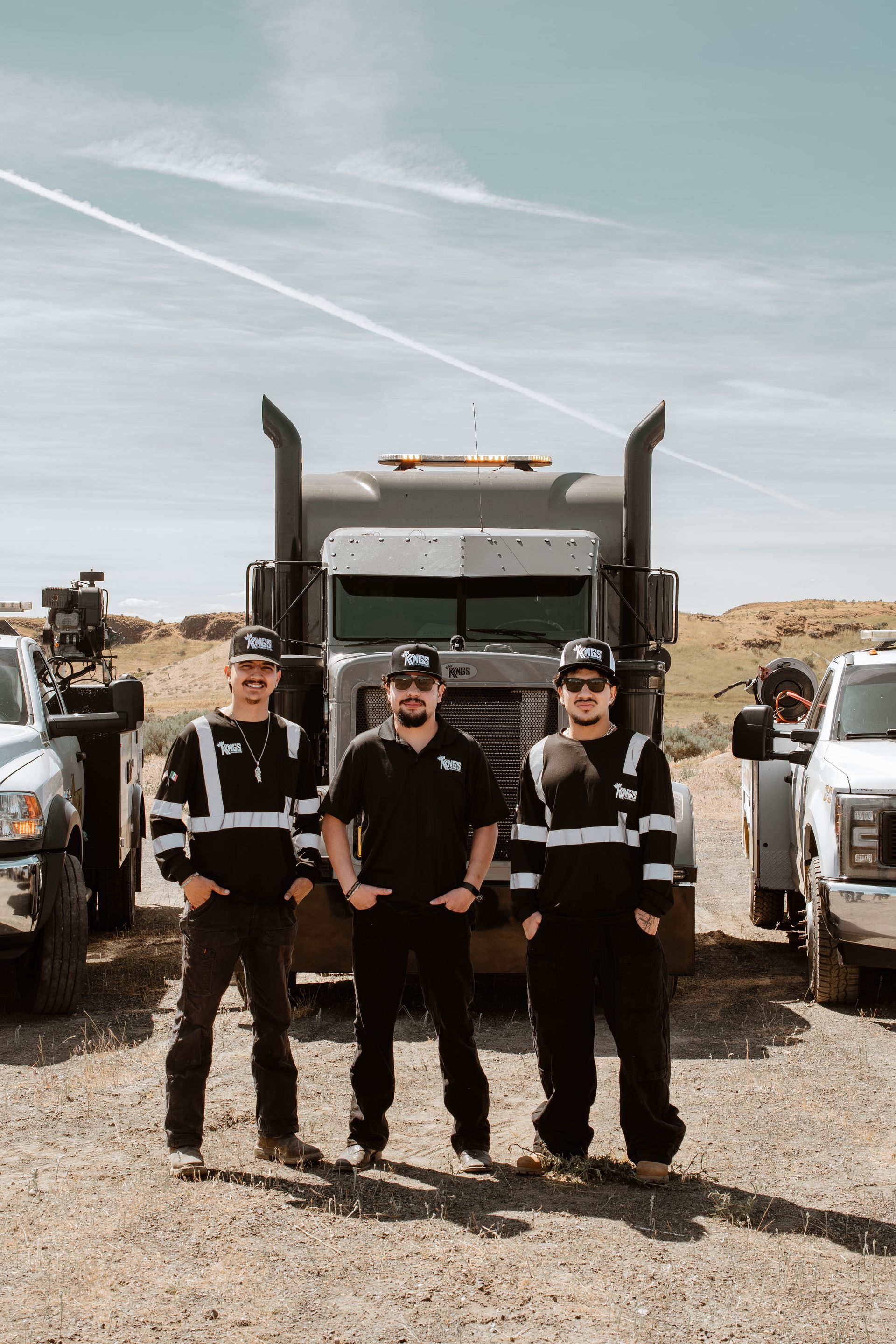 Three people in dark work gear standing in front of a large truck in a desert setting.