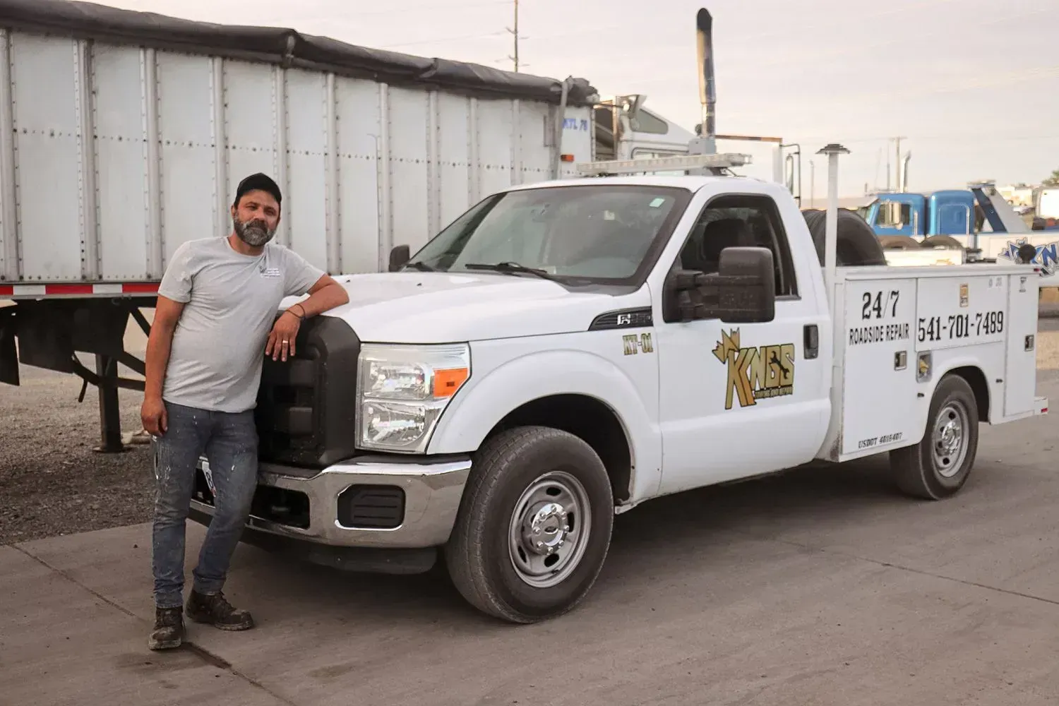 A man stands beside a white service truck in an outdoor lot, with a large trailer visible in the background.