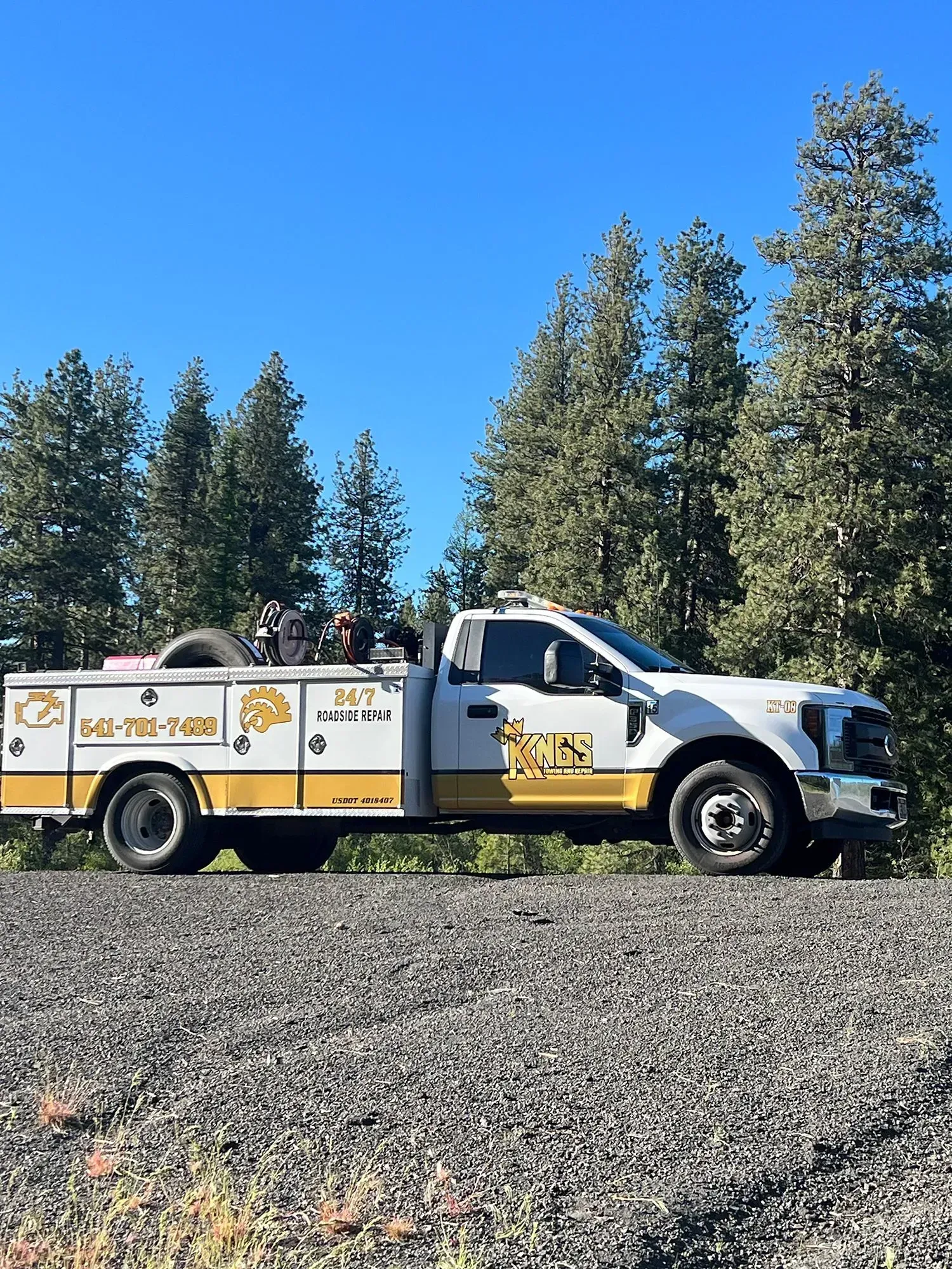 A white and tan service truck parked on a gravel road with a forest backdrop under a clear blue sky.