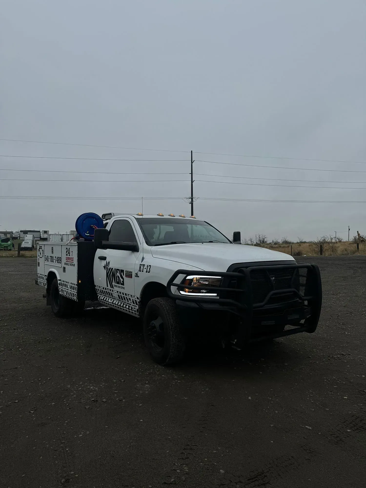 A white utility truck with a black brush guard and decals parked on a gravel lot under an overcast sky.