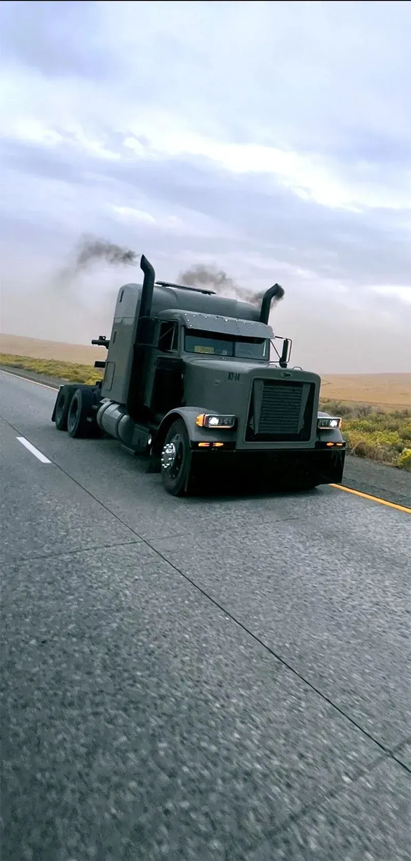 A matte dark grey semi-truck with smoke emitting from its stacks travels down a highway through an open landscape.