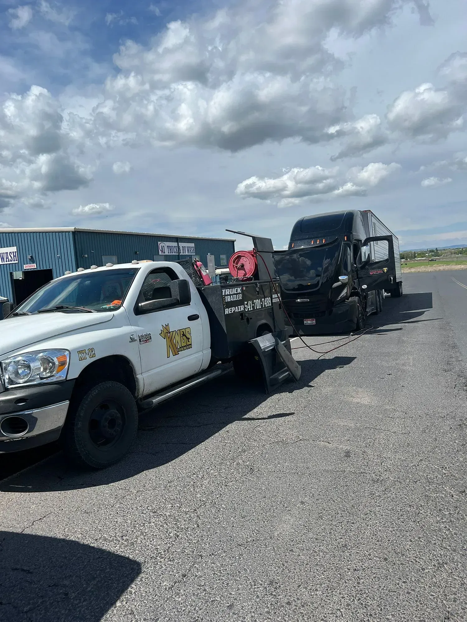 A white service truck is towing a black semi-truck on a gravel surface under a partly cloudy sky.