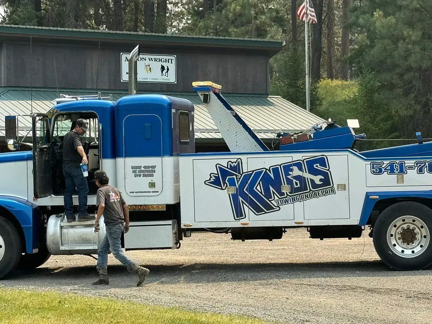 Two people stand by a blue and white KNGS towing truck parked in a gravel lot in front of a building with an American flag.
