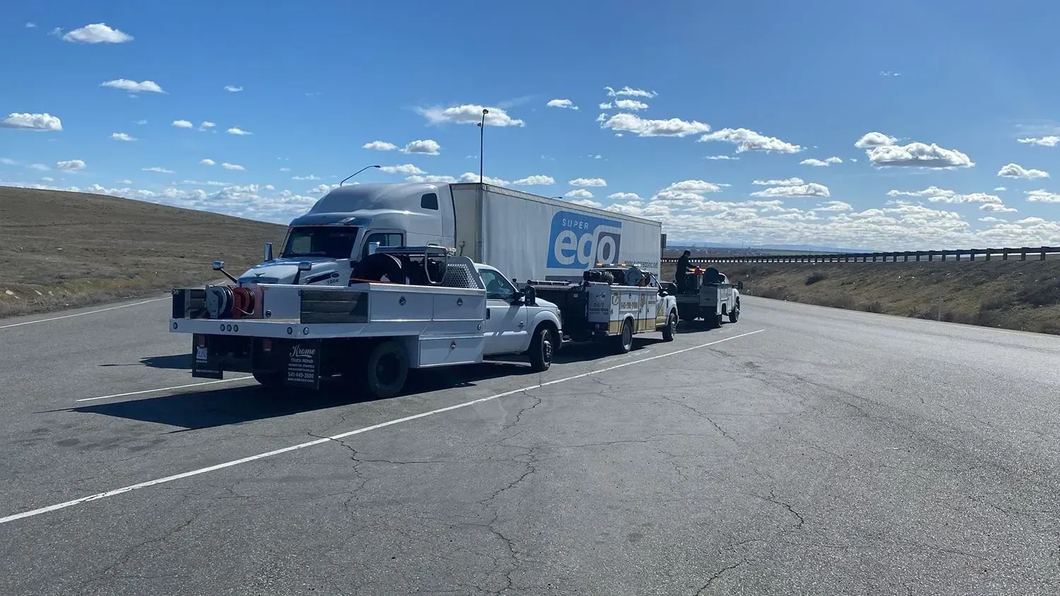 White trucks and a large trailer parked on the side of a highway under a clear blue sky.