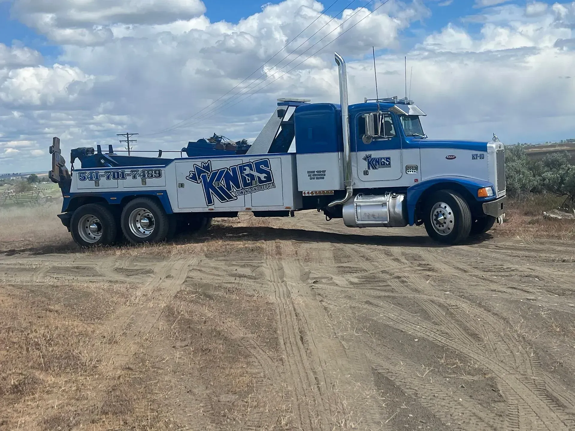 A blue and white heavy-duty tow truck driving on a dusty, unpaved field under a cloudy sky.