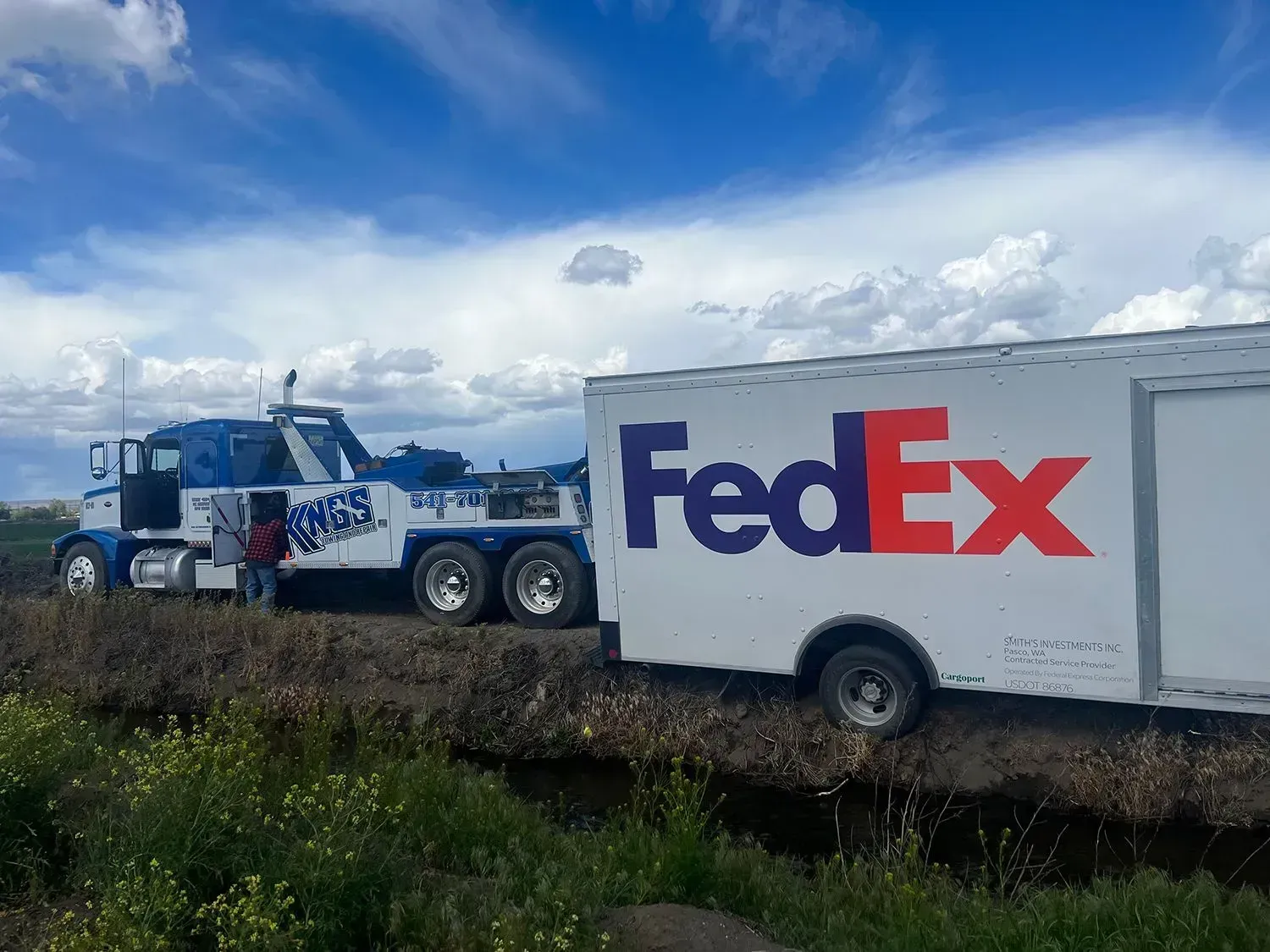 A blue tow truck pulling a white FedEx trailer stuck in a muddy field under a cloudy sky.