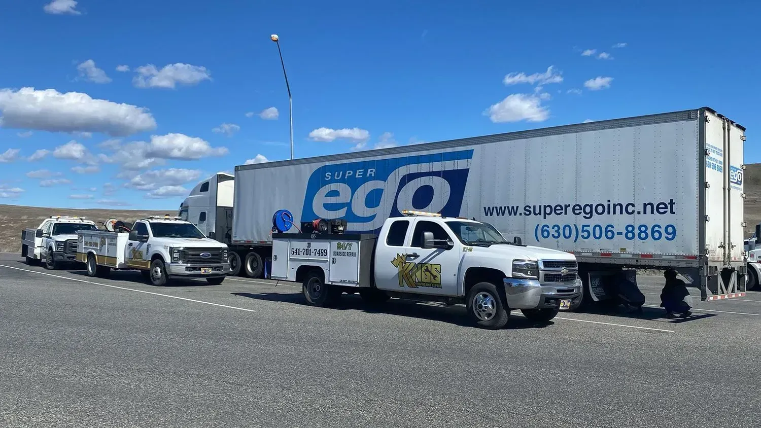 White Ego-branded semi-truck and trailer parked outdoors next to two service trucks on a gravel lot under a blue sky.